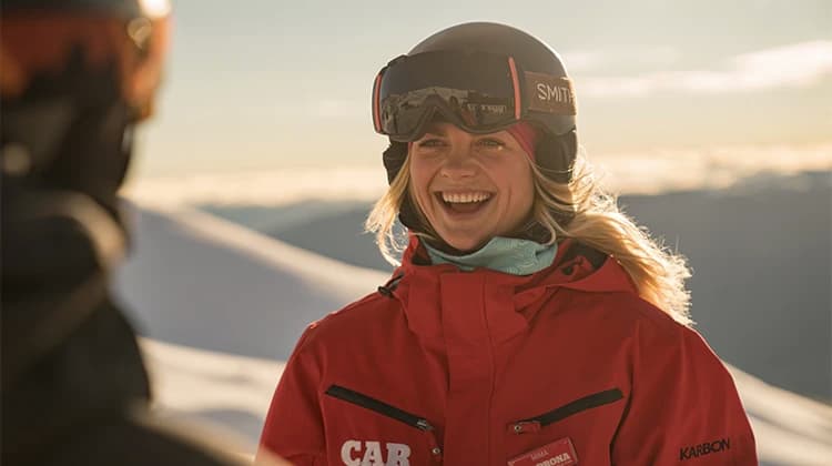 Close up shot of a woman smiling while standing outside with snowy mountains in the background. She is wearing a red ski jacket branded 'Cardrona' and a a black ski helmet with goggles branded 'Smith' on the helmet. 