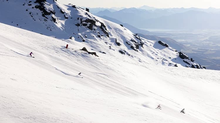 The image shows two skiers and two snowboarders carving down a snow-covered slope at Cardrona Alpine Resort, with rocky outcrops nearby. In the distance, a stunning view of Lake Wanaka and layers of mountain ranges stretch across the horizon, creating a breathtaking alpine backdrop.