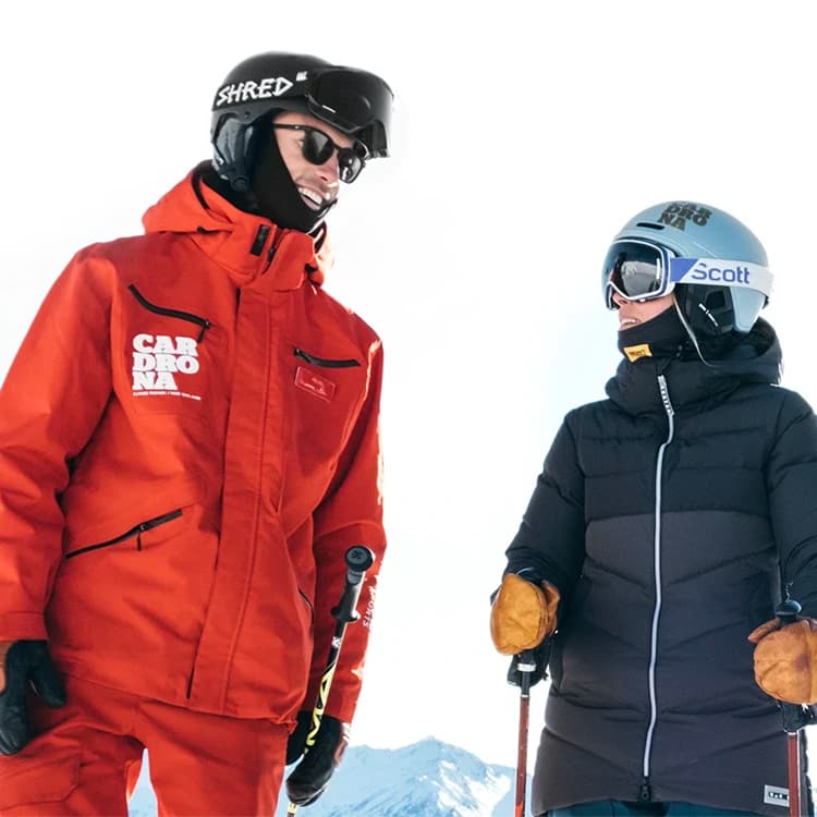 Two skiers stand together on a mountain peak, sharing a laugh. The skier on the left is dressed in a bright red jacket with "Cardrona" branding. The skier on the right, wearing a dark puffy jacket and a helmet with a "Scott" label, listens intently while smiling. 