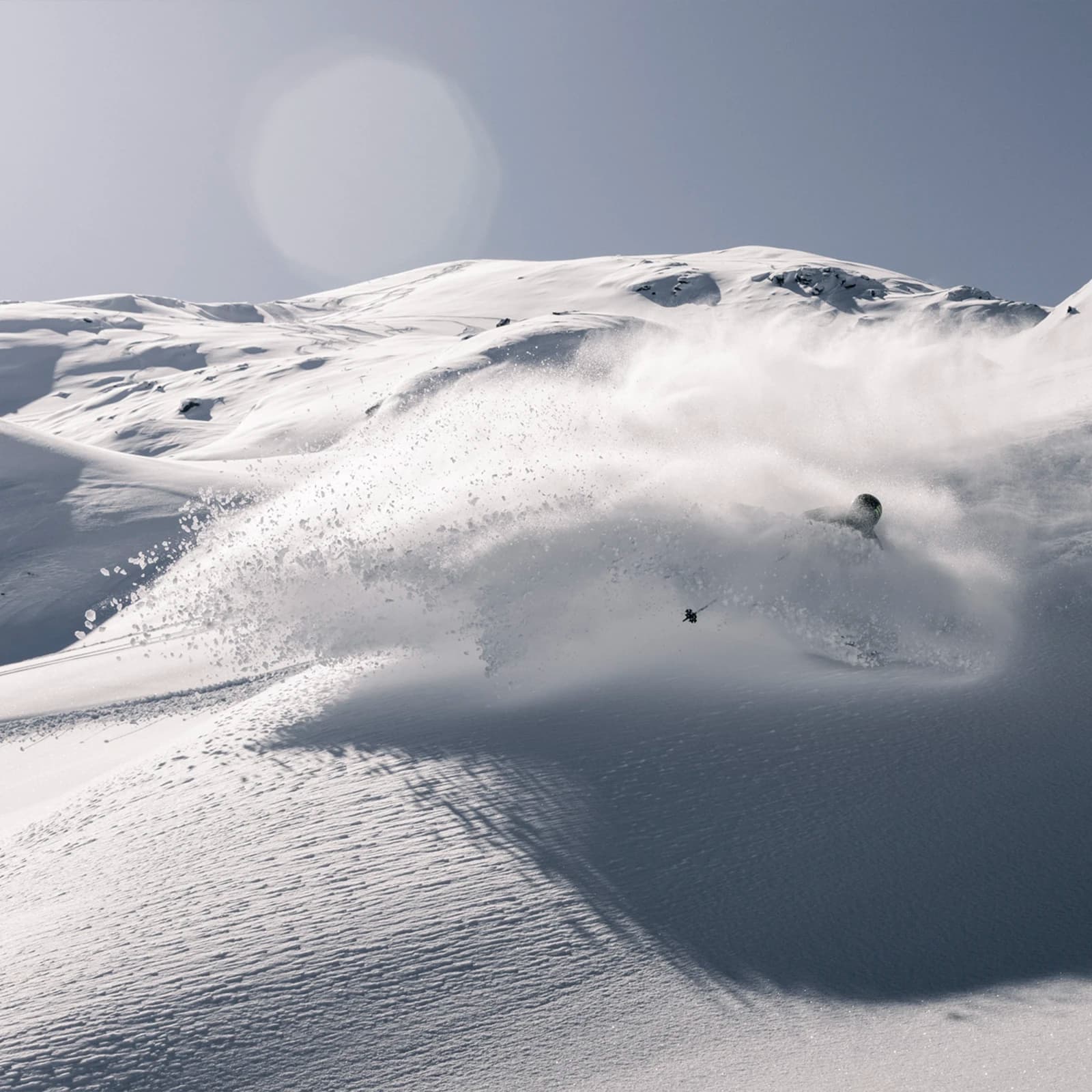 A skier carves through deep powder in Soho Basin at Cardrona Alpine Resort, sending a spray of snow into the air. The untouched snowy slope glistens under clear skies, with the sun casting soft shadows across the pristine landscape, highlighting the skier’s graceful movement.