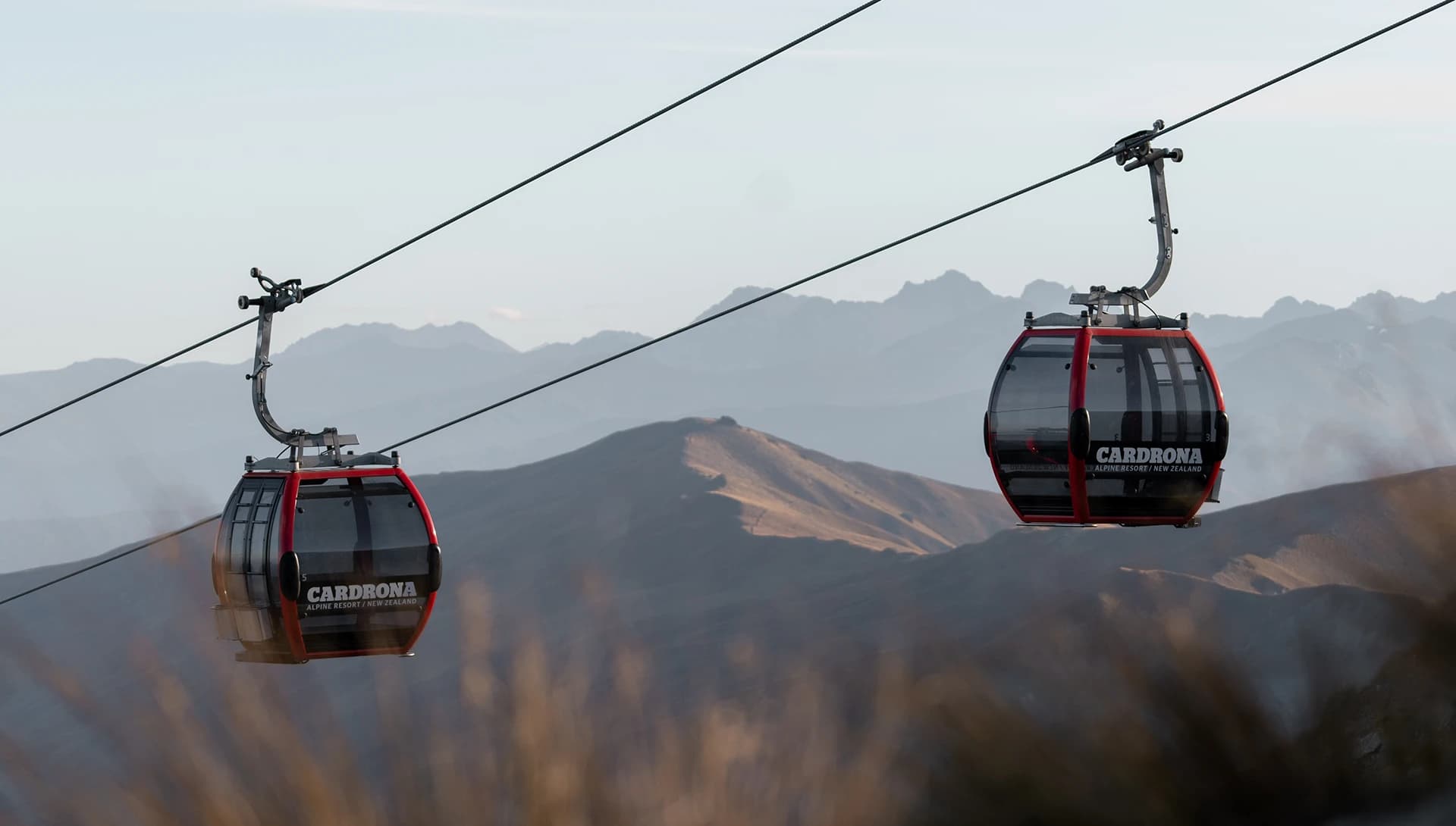 Two red gondolas glide along cables above a rugged alpine landscape at Cardrona Alpine Resort in New Zealand. The distant mountain ranges create a scenic backdrop under a clear sky.