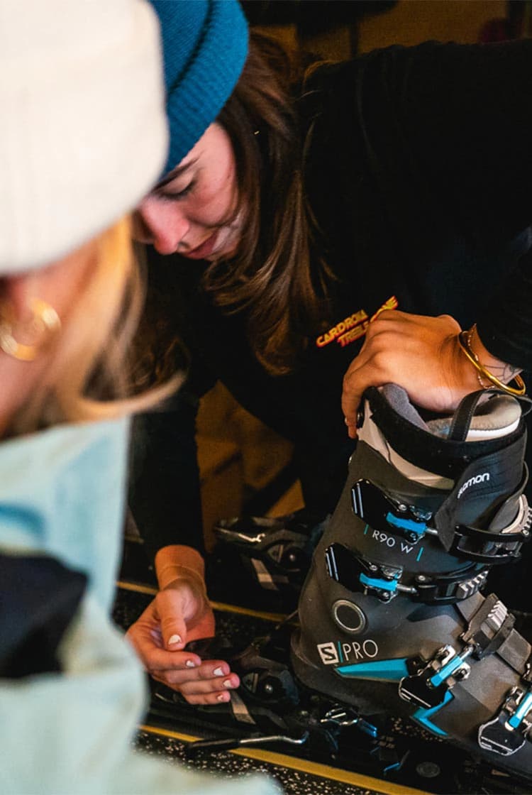 An indoor scene at a ski rental shop with a female technician adjusting ski boots for a customer. The technician, wearing a black long-sleeve shirt with the "Cardrona Rentals" logo, is focused on fitting a black and blue Salomon ski boot onto the bindings. The customer, partially visible, is wearing a light blue jacket and a white beanie.