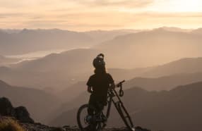 A mountain biker stands with their bike at sunset, overlooking the expansive Cardrona mountain range and valleys below.