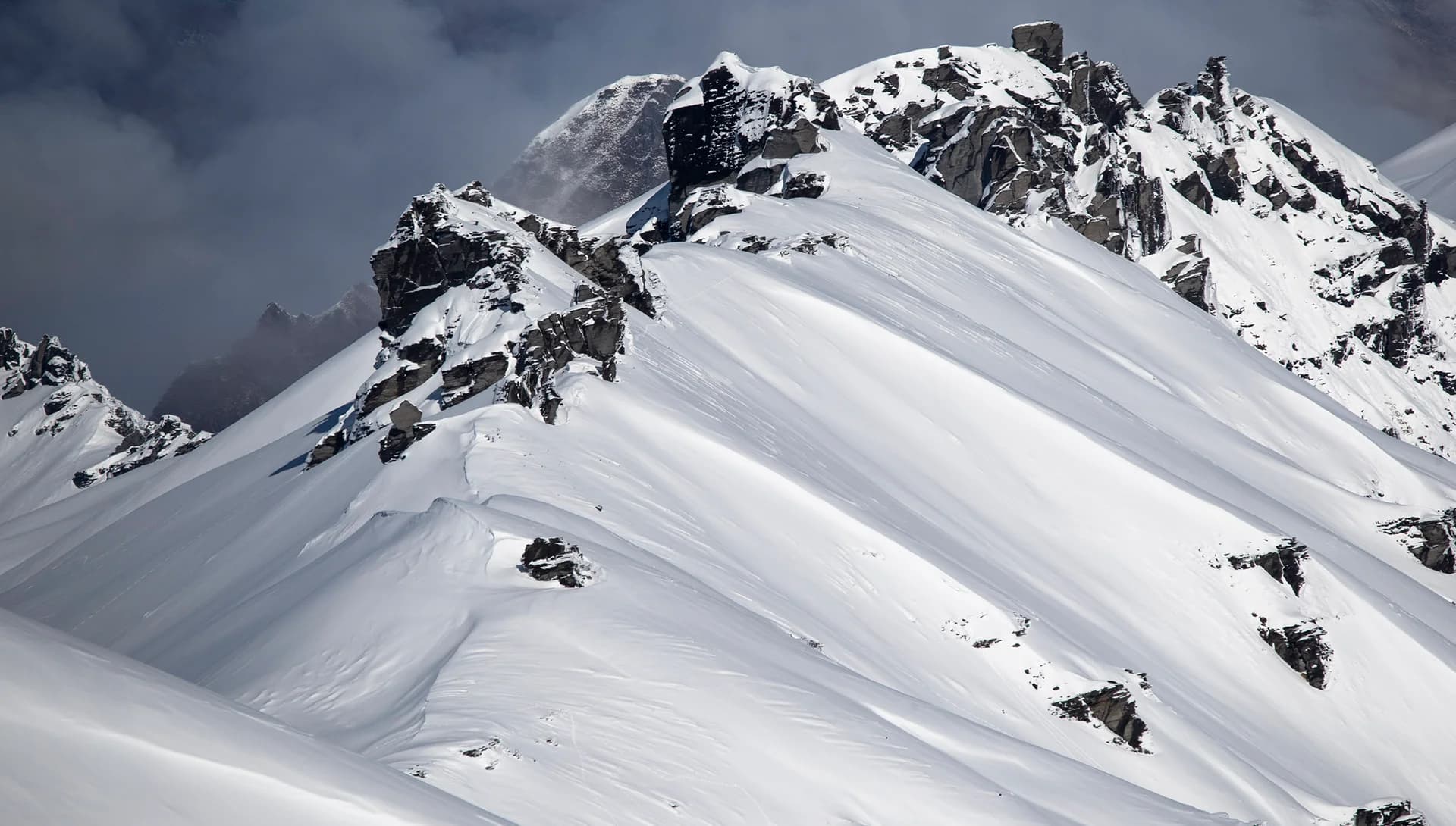 A dramatic snow-covered mountain ridge with sharp, jagged rock formations breaking through the smooth, pristine snow. The contrast between the rugged, dark rocks and the bright white snow creates a striking visual. In the background, thick clouds or mist envelop parts of the mountains, adding to the sense of isolation and raw natural beauty. The untouched snow and steep terrain suggest a remote, backcountry setting, emphasising the challenge and allure of alpine environments for adventurers and winter sports enthusiasts.