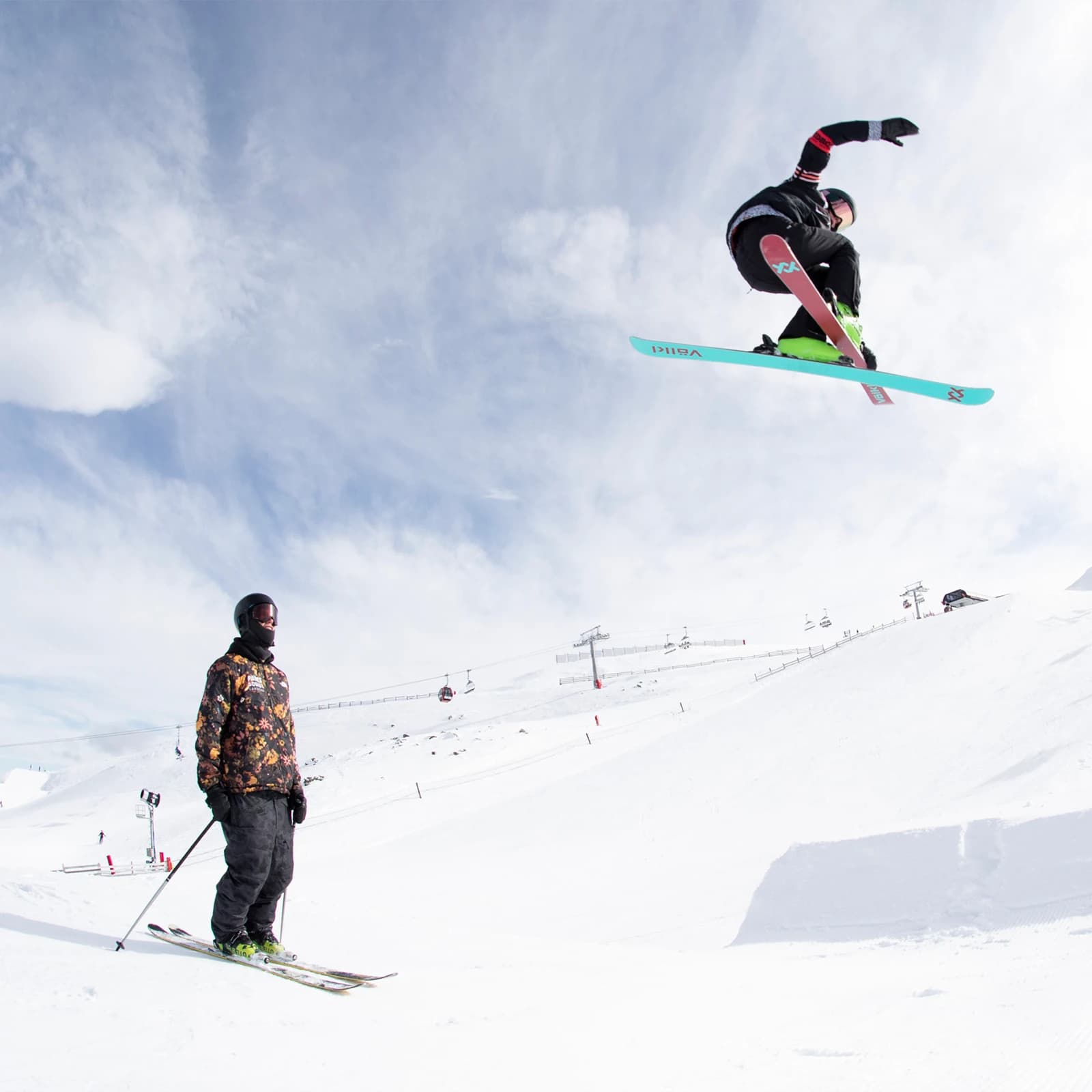 A skier performing a high-flying trick, grabbing the skis mid-air, with vibrant blue skies and scattered clouds in the background. Another skier, dressed in a colourful jacket and dark pants, watches from the slope below. The backdrop includes a snow park, with visible ski lifts and other skiers in the distance, adding to the energy of the scene. 