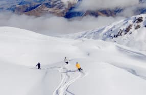 Three skiers glide down the snowy slopes of Soho Basin at Cardrona Alpine Resort, surrounded by vast, untouched powder and majestic mountain peaks. The scene is set against a backdrop of cloud-covered ridges and snow-capped peaks, offering a pristine and expansive winter landscape.