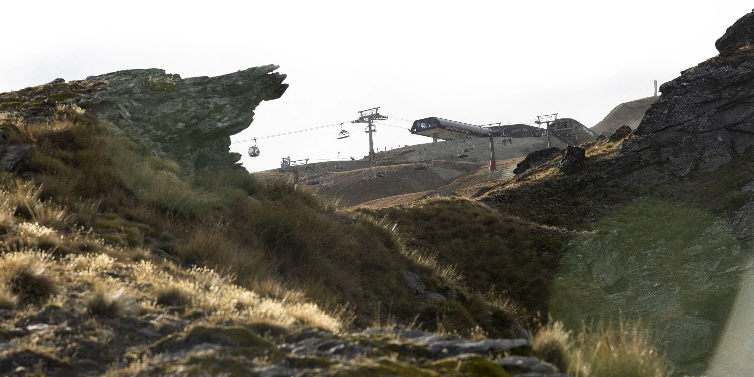 A rugged mountainous landscape with steep rock formations and grassy slopes during summer. A chair lift, with gondolas and chairlifts, is visible in the background. 