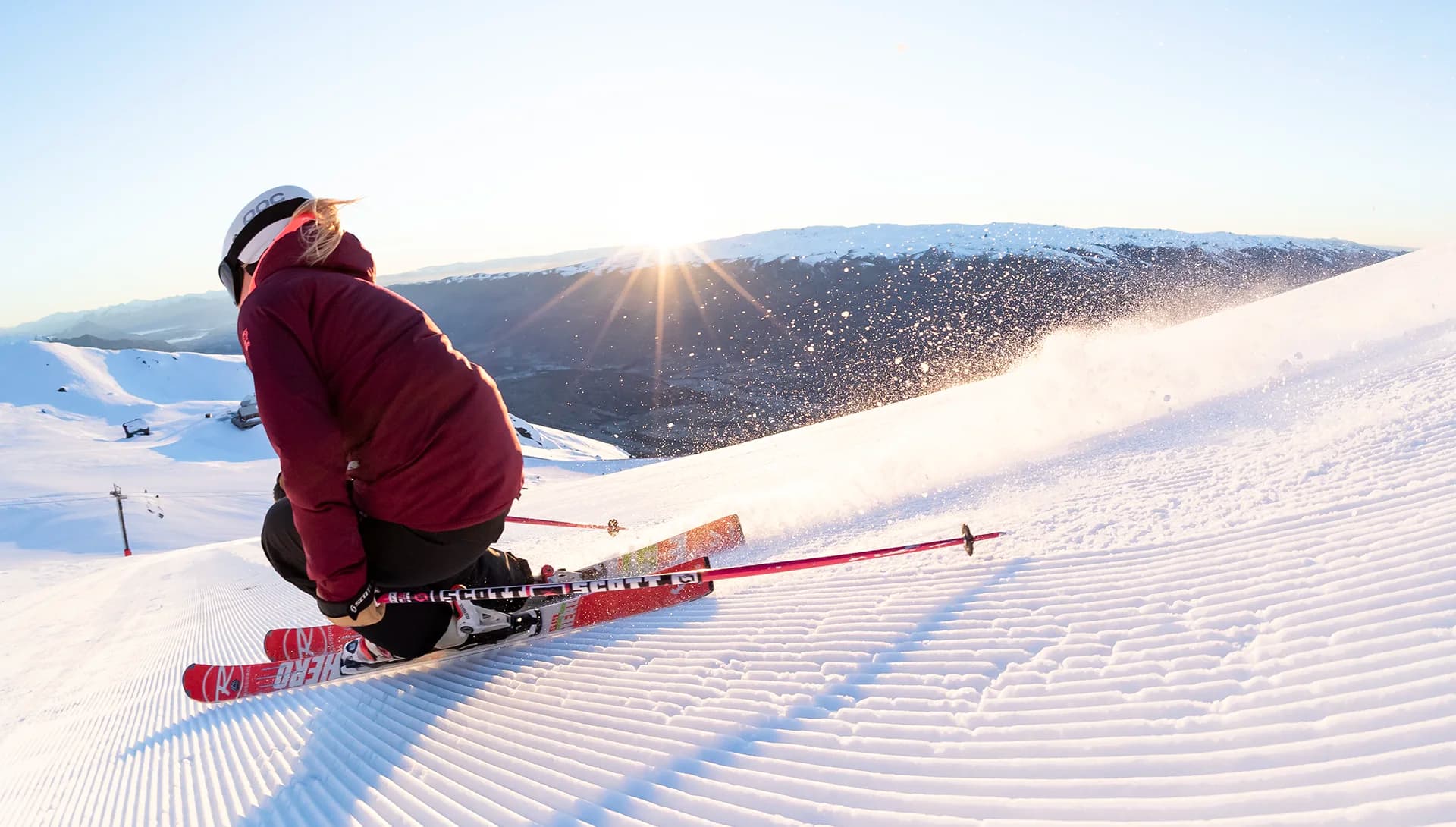 A skier carves through the freshly groomed snow at Cardrona, with the sun rising over the distant mountains, casting a golden light on the slopes as snow sprays up behind them.