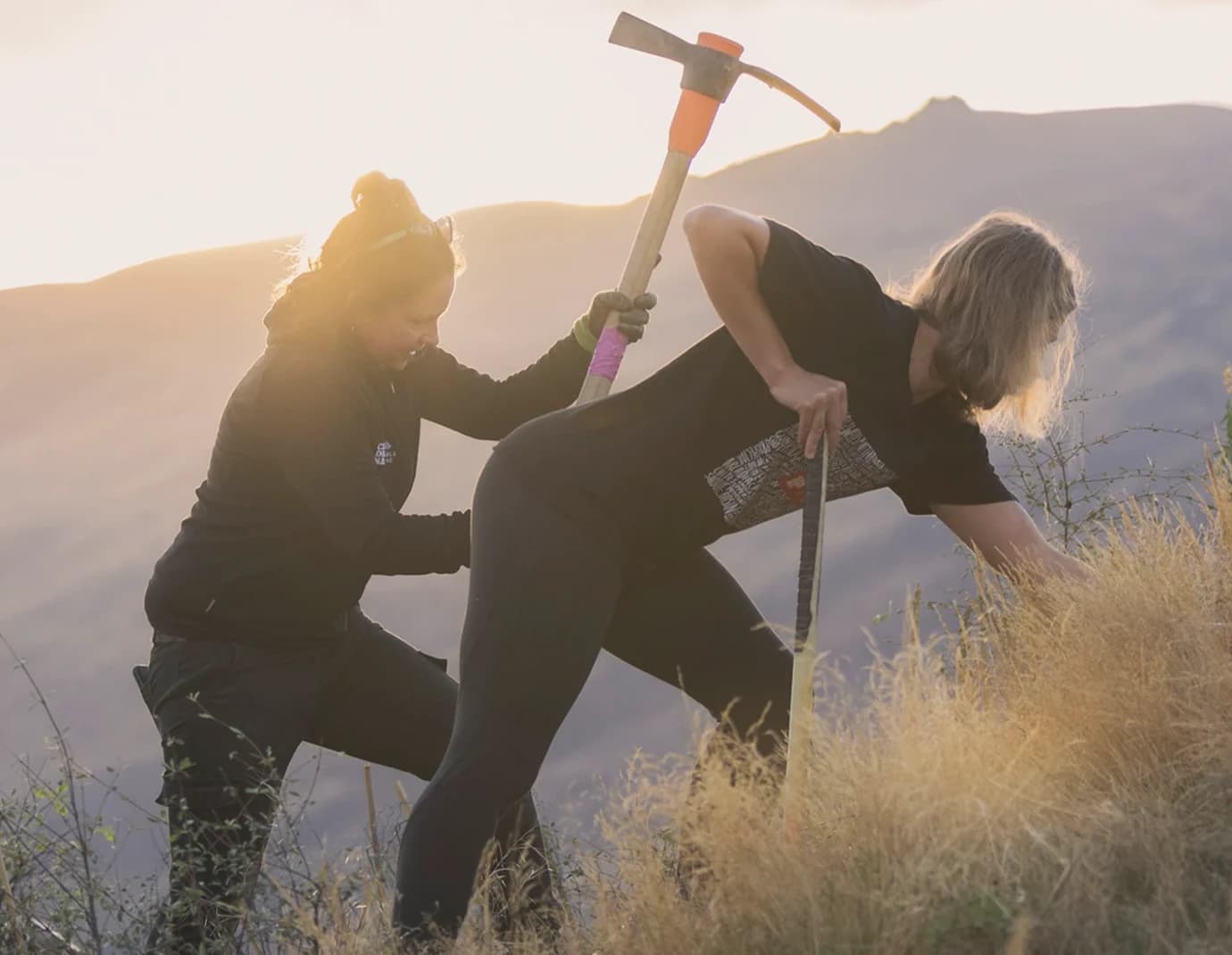 Two people work together on a hillside, one holding a pickaxe and the other using a spade to dig into the dry grass, with soft sunlight illuminating the mountainous background.