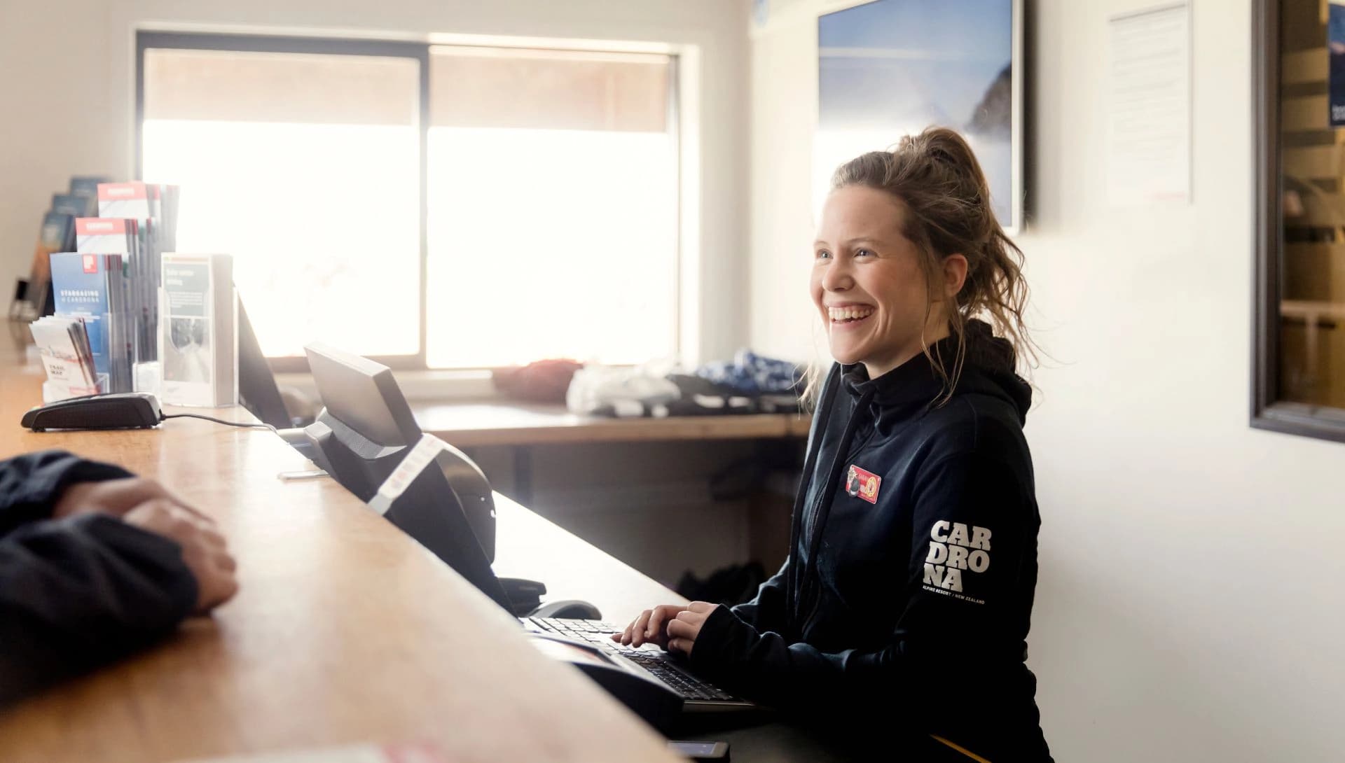 A smiling staff member at Cardrona Alpine Resort assists a guest from behind a reception desk, creating a warm and welcoming atmosphere in a brightly lit space.