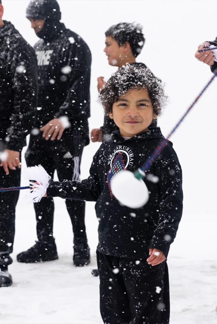 A young child dressed in black smiles while playing with traditional poi in the snow, as others participate in the background, with snow gently falling around them.