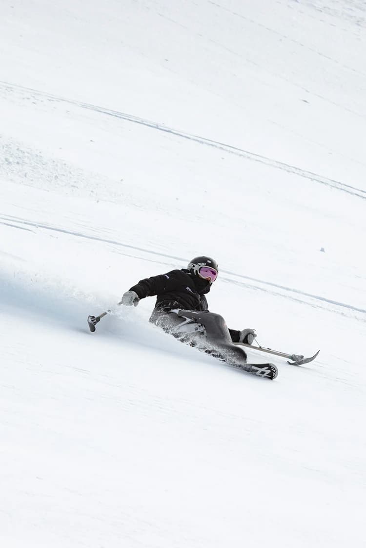 A skier in adaptive equipment carves through fresh snow at Treble Cone Ski Field, leaving a trail of powder on the steep, snowy slope.