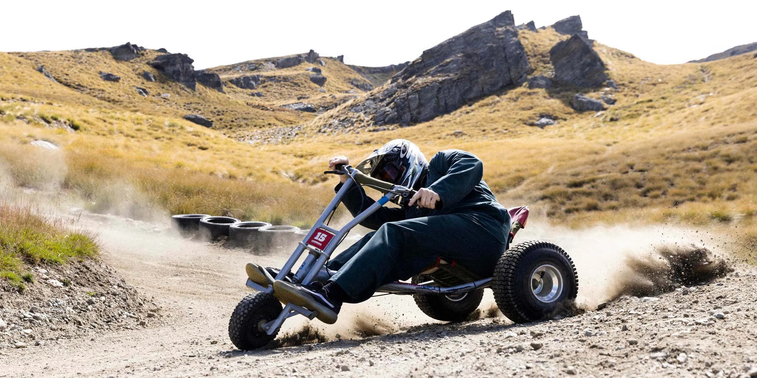 The image captures an intense moment of gravity carting at Cardrona Alpine Resort. A rider in a dark green outfit and helmet leans into a turn on a dirt track, kicking up dust as they navigate the course. The rugged, mountainous terrain with grassy hills and scattered rocks provides a dramatic backdrop to the action. The rider’s focused posture and the dirt flying from the wheels highlight the adrenaline-pumping excitement of mountain carting at the resort.