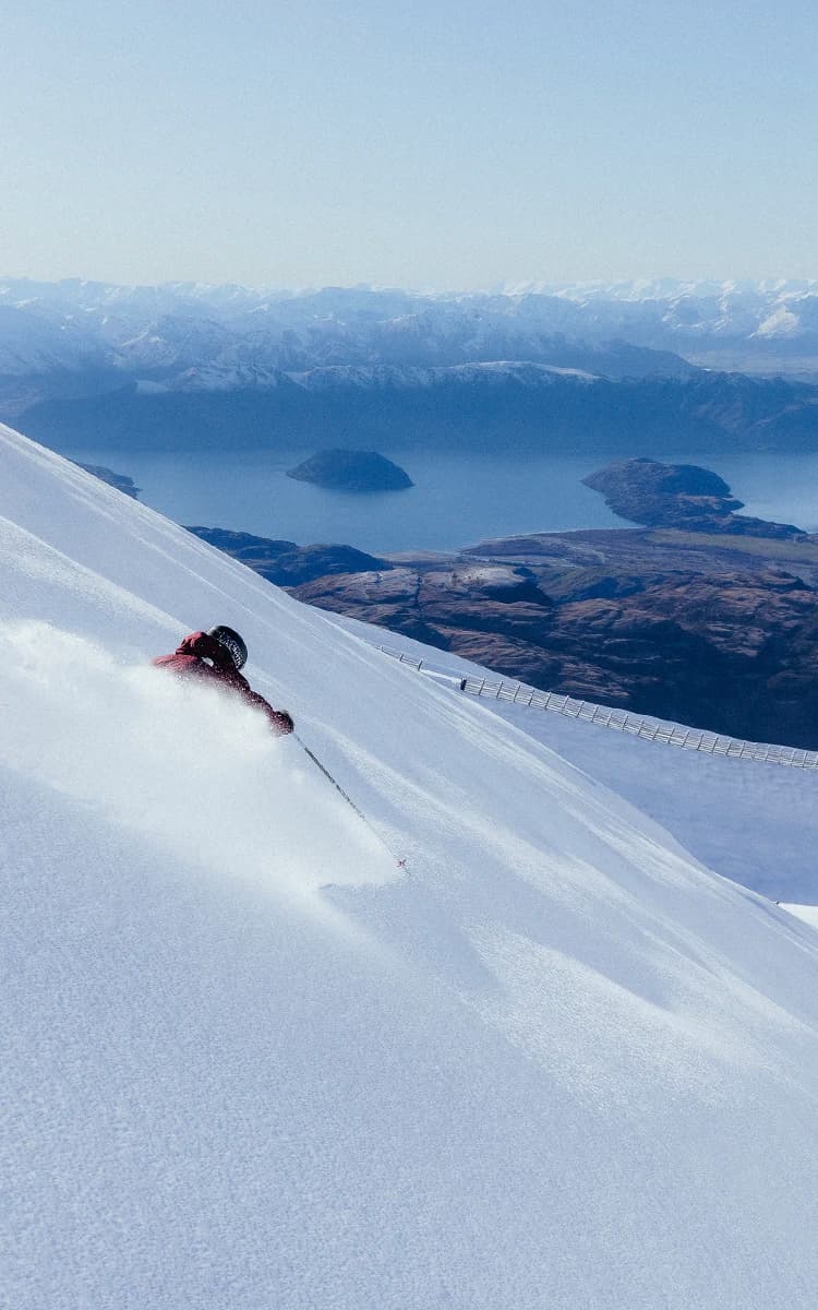 A skier carves down a steep, snowy slope with powder flying, while a vast lake and snow-capped mountains stretch across the background.