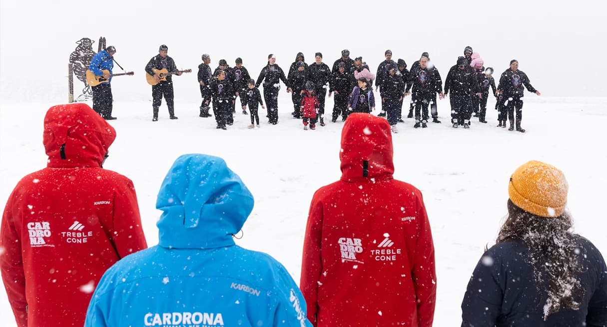 A snowy outdoor scene with a group of people standing in a circle. In the foreground, three individuals with their backs to the camera are wearing bright-coloired jackets (two red and one blue) branded with the logos "Cardrona" and "Treble Cone." In the distance, a larger group of people, dressed mostly in black, are gathered in the snow, some playing guitars. The scene appears to be a ceremony or cultural gathering, possibly a welcome or performance. Snow is falling, adding to the wintry atmosphere, and everyone seems engaged in the event despite the cold.