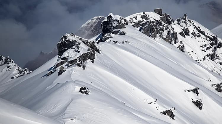 A dramatic snow-covered mountain ridge with sharp, jagged rock formations breaking through the smooth, pristine snow. The contrast between the rugged, dark rocks and the bright white snow creates a striking visual. In the background, thick clouds or mist envelop parts of the mountains, adding to the sense of isolation and raw natural beauty. The untouched snow and steep terrain suggest a remote, backcountry setting, emphasising the challenge and allure of alpine environments for adventurers and winter sports enthusiasts.