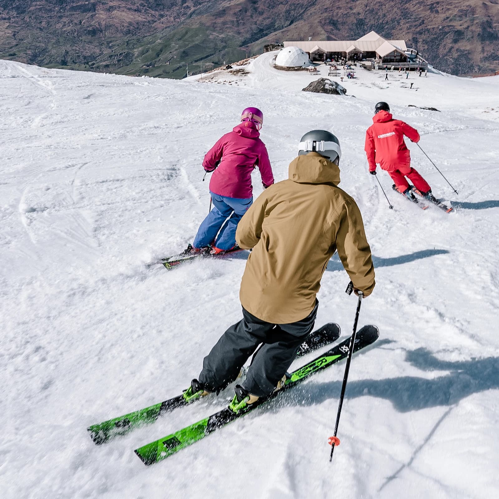 Three skiers, dressed in vibrant gear, glide down a snowy slope at Cardrona, following an instructor in red. A lodge is visible in the background, with scenic mountain views surrounding the area.