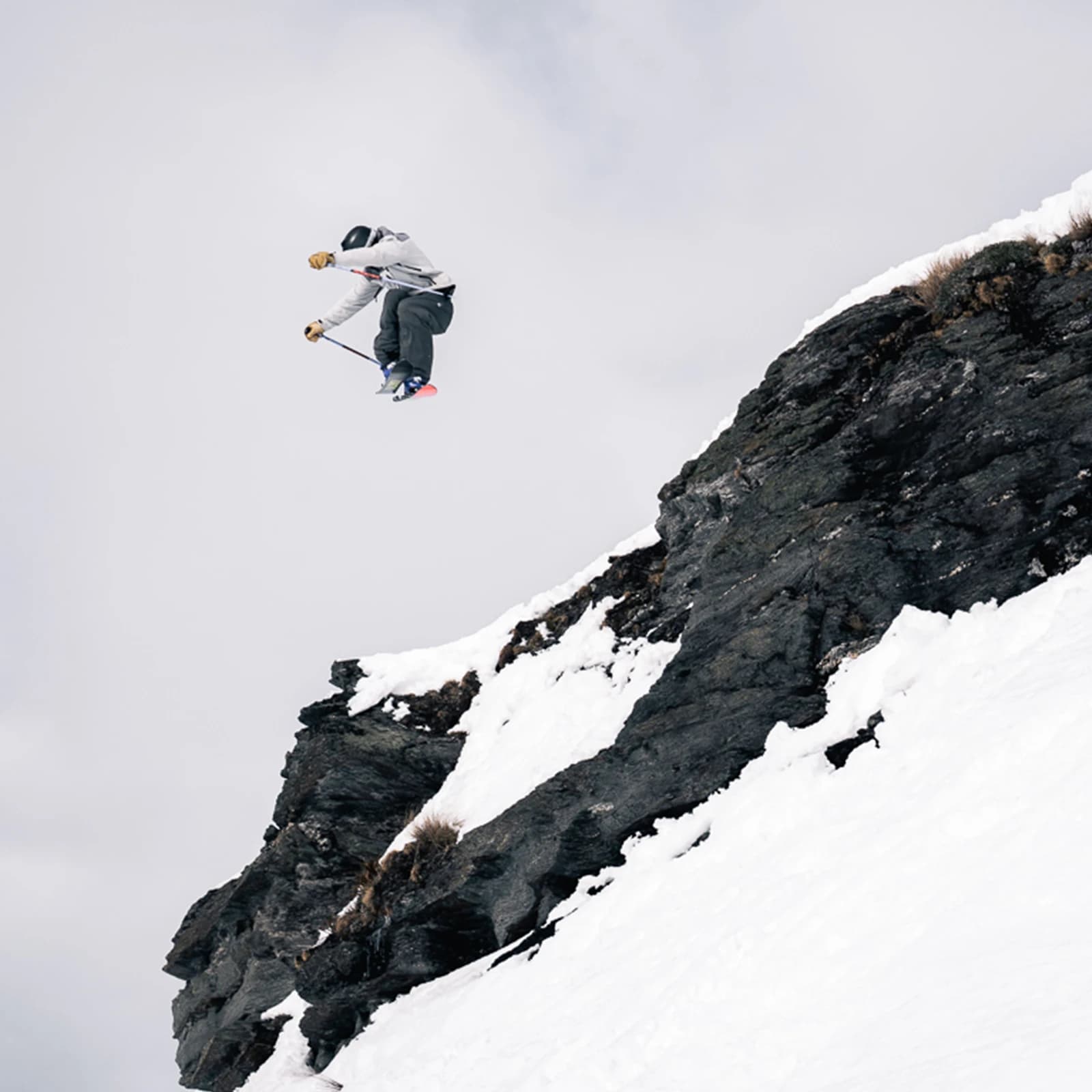 A skier performs a jump from a rocky outcrop in a snowy landscape. The skier in the air is mid-jump. 