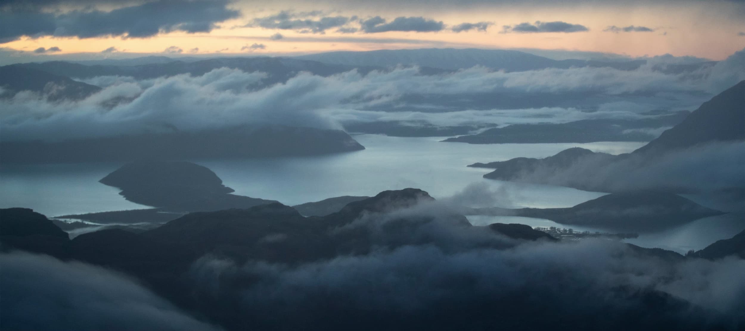 A stunning aerial view of mist-covered mountains and lakes at dusk, with layers of clouds gently floating over the water and peaks, creating a serene and mystical landscape.