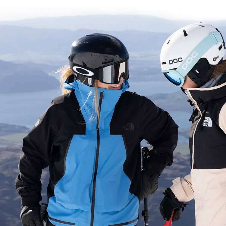 Two skiers wearing helmets and goggles stand on a snowy mountain, chatting with a scenic view of a lake and mountains in the background.