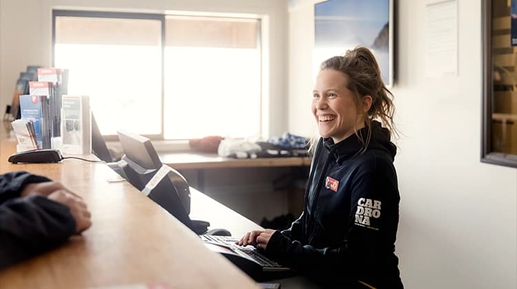 A smiling staff member at Cardrona Alpine Resort assists a guest from behind a reception desk, creating a warm and welcoming atmosphere in a brightly lit space.