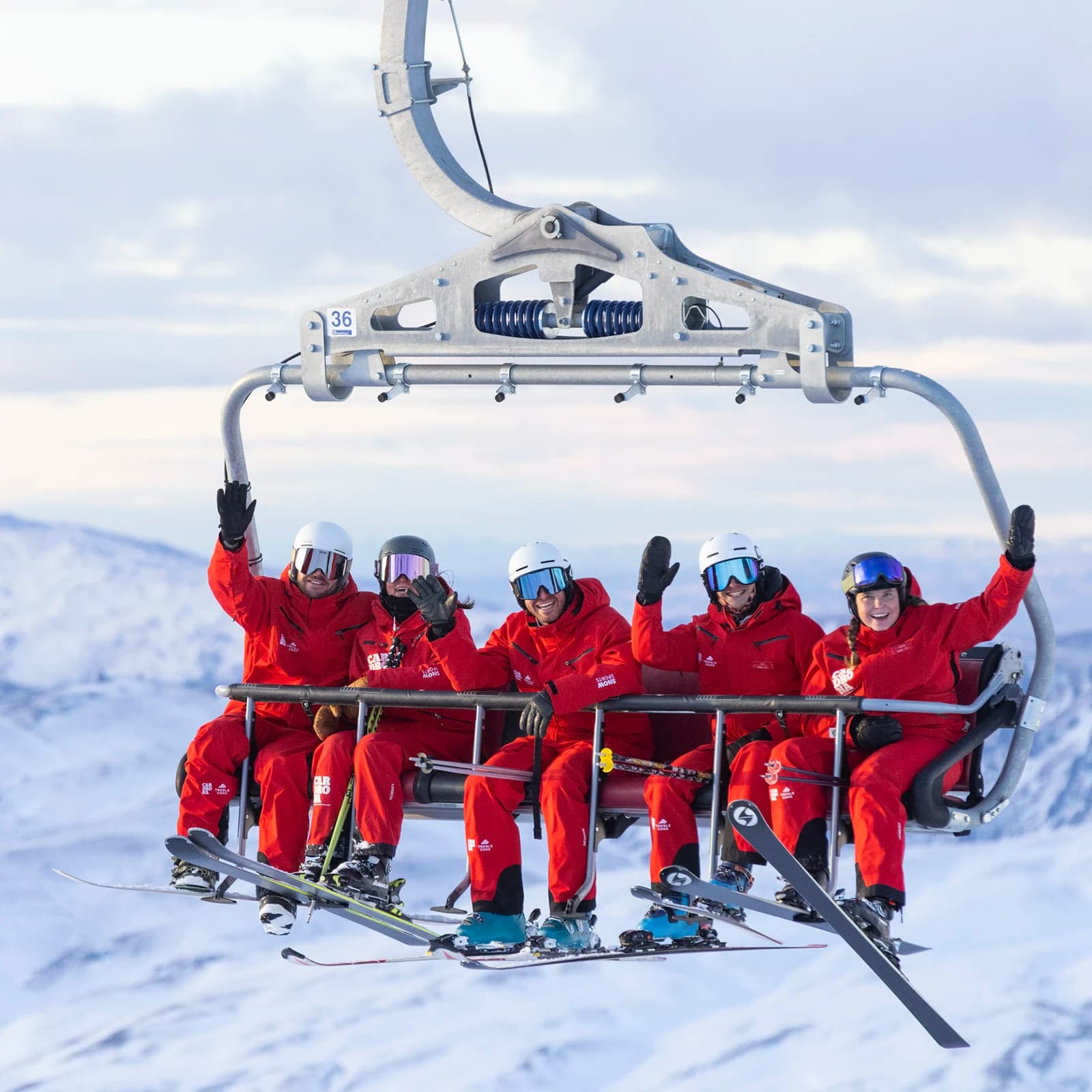 Five skiers in red gear ride a chairlift, waving as they ascend over a snowy mountain landscape. Clear skies and distant peaks frame the scene.