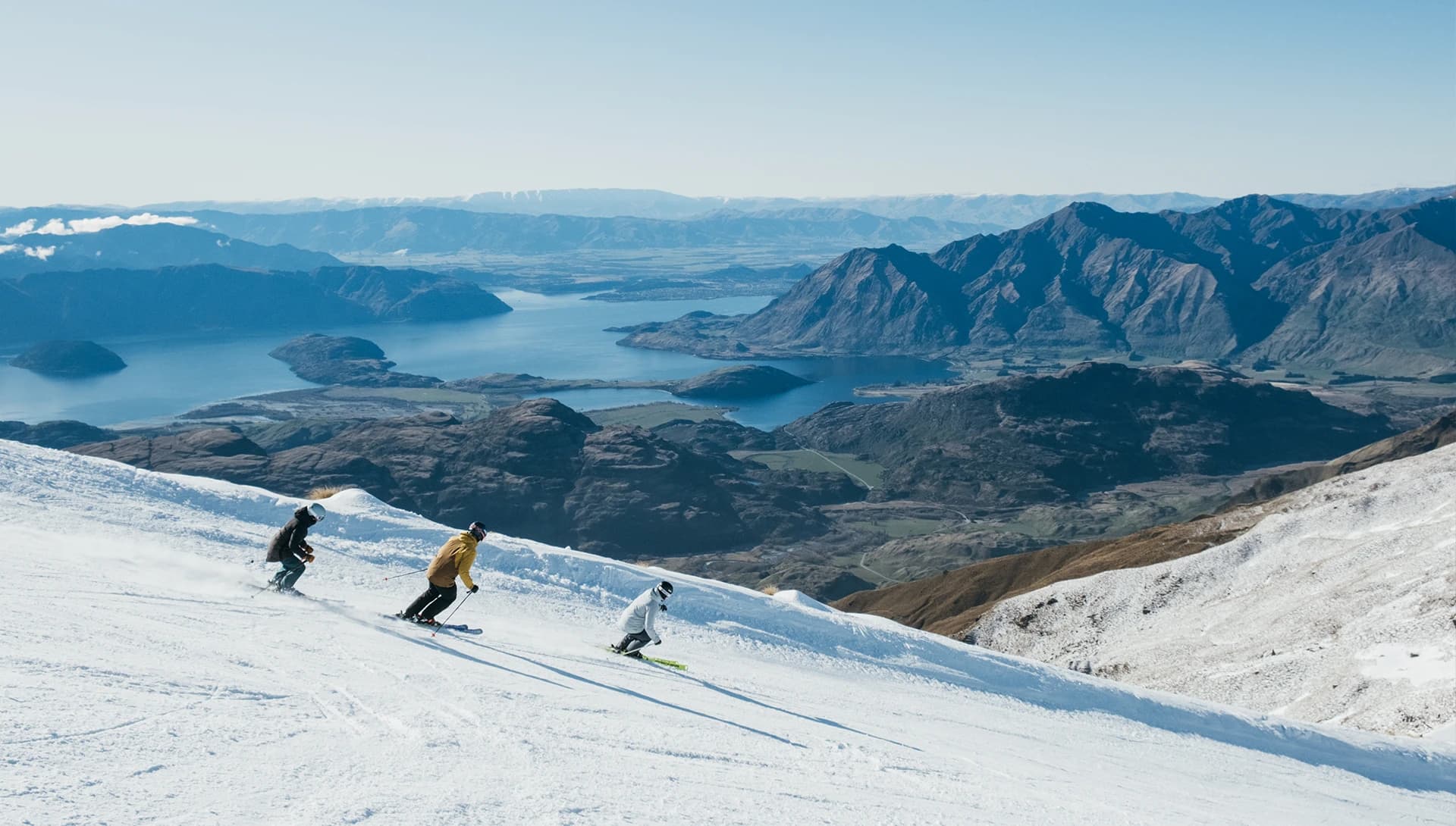 Three skiers descending a snowy slope on a clear day with a vast mountainous landscape in the background. Below the mountain range, a large blue lake is surrounded by green valleys. 