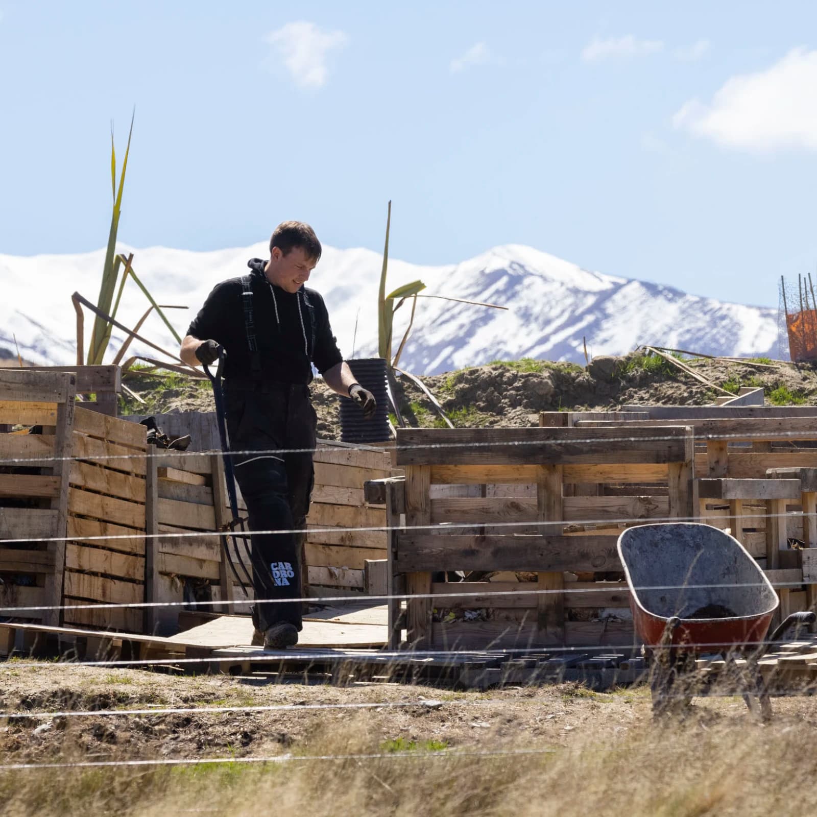 A person working outdoors near a composting site. The individual is dressed in dark clothing with "Cardrona" visible on the leg, indicating a possible affiliation with the Cardrona ski area. 