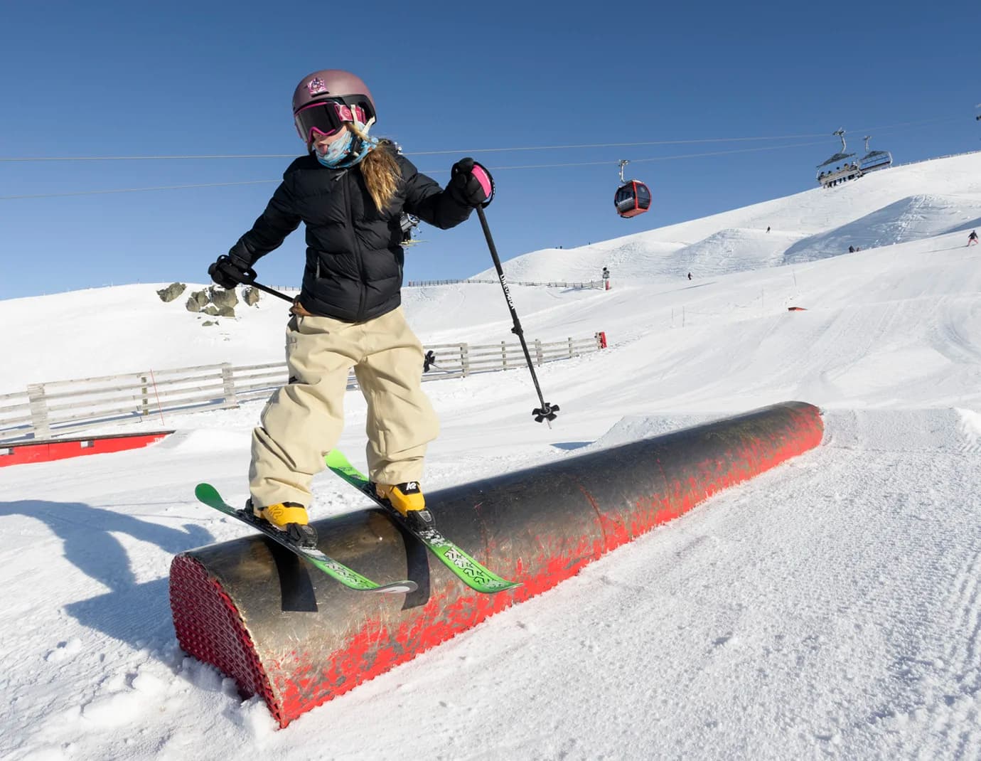 A young skier balances on a rail during a freestyle session in the Cardrona park. Wearing a helmet and goggles, the skier confidently glides over the obstacle with ski poles in hand.