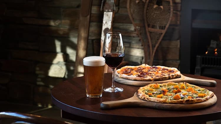 Two wood-fired pizzas on boards, a glass of red wine and a pint of beer sit on a table in a lounge area with a lit fireplace in the background.