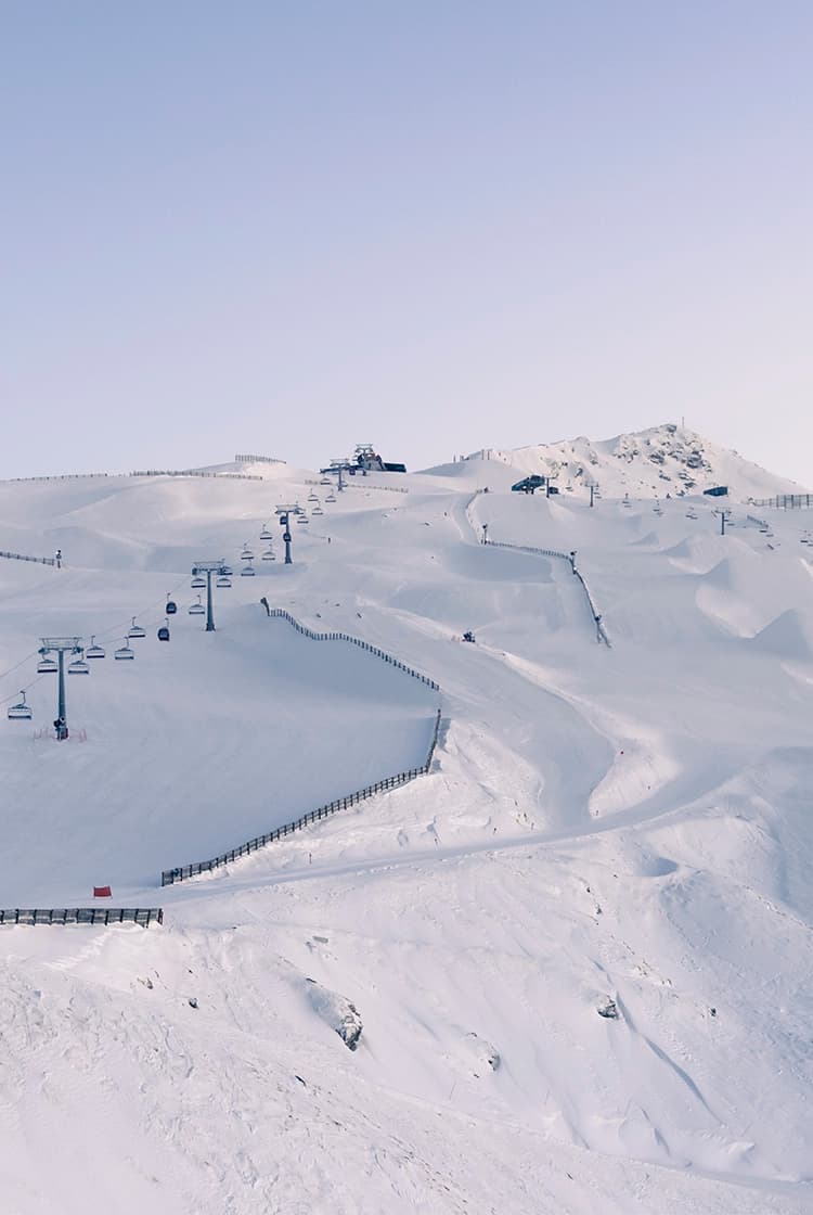 A vast, snow-covered ski resort at Cardrona with smooth, groomed slopes and ski lifts ascending the mountain. The clear blue sky and expansive terrain create a perfect winter sports scene, with skiers and snowboarders visible in the distance.