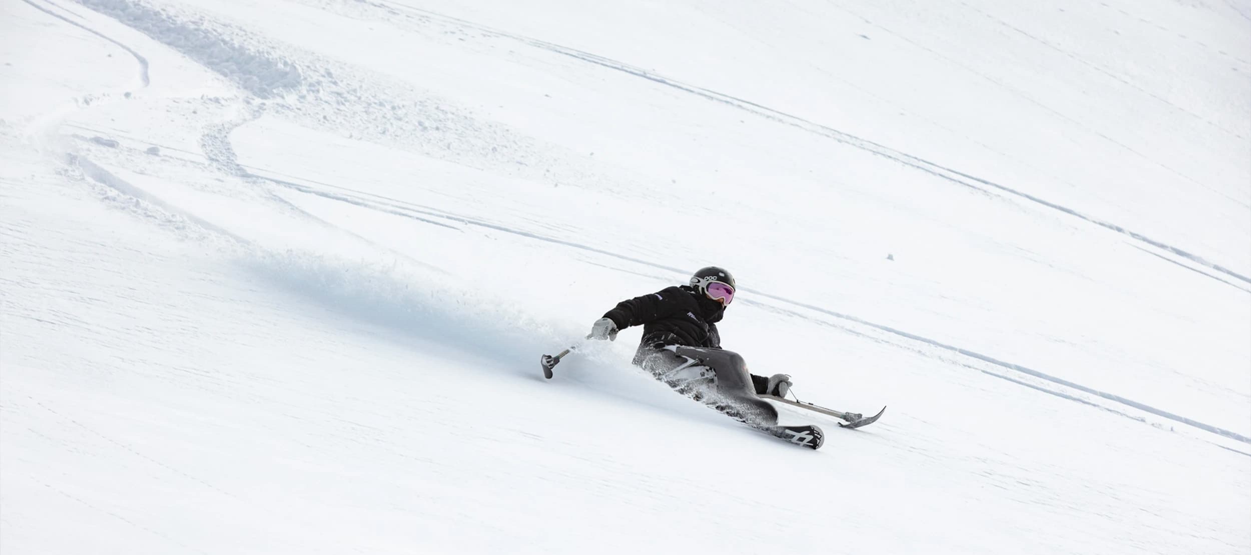 A skier in adaptive equipment carves through fresh snow at Treble Cone Ski Field, leaving a trail of powder on the steep, snowy slope.