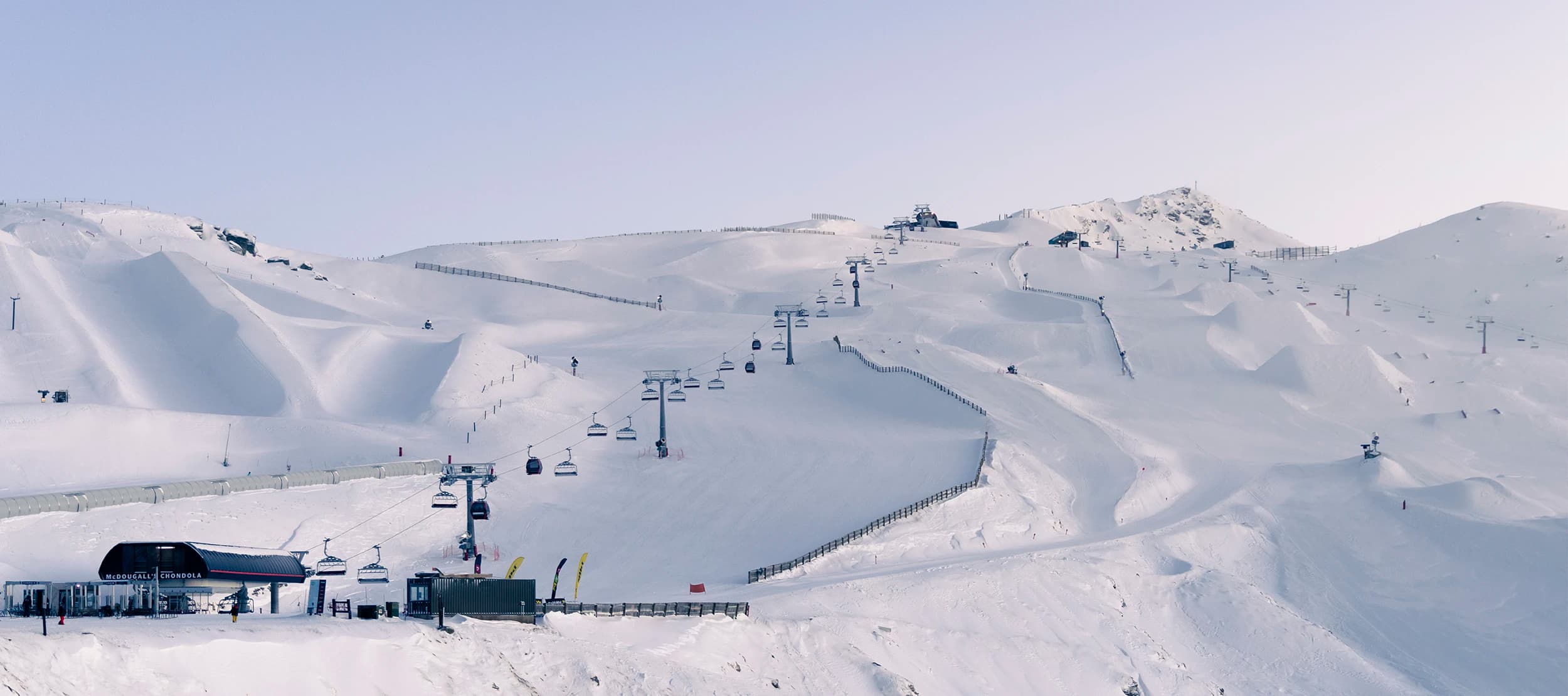 A vast, snow-covered ski resort at Cardrona with smooth, groomed slopes and ski lifts ascending the mountain. The clear blue sky and expansive terrain create a perfect winter sports scene, with skiers and snowboarders visible in the distance.