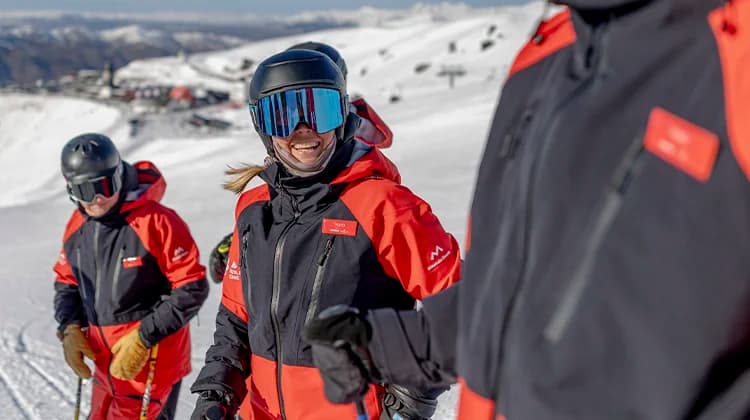 A group of skiers in red and black gear, including helmets and goggles, stand together smiling on a sunny, snow-covered mountain with a resort and chairlifts visible in the background.