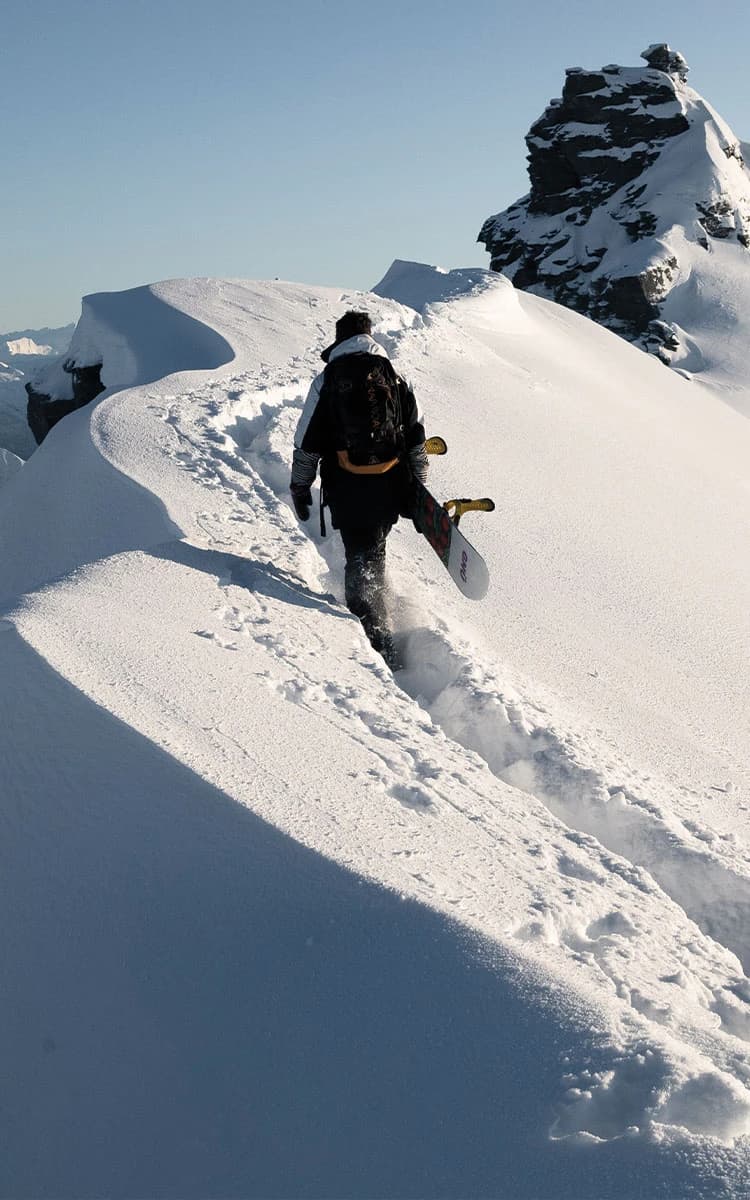 A snowboarder hikes along a narrow, snow-covered mountain ridge with breathtaking views of rugged peaks and deep valleys in the distance under a clear blue sky.