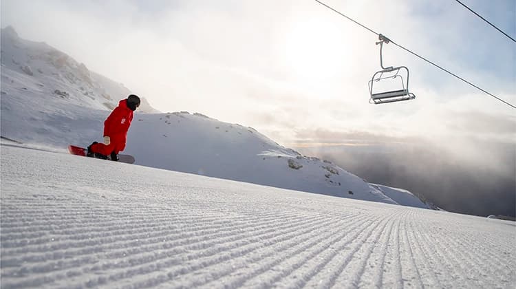 A lone snowboarder in a red jacket riding a freshly groomed snow-covered slope. The grooves in the snow lead the viewer's eye toward the distant mountains and the bright sun peeking through light clouds. A single ski lift chair hovers overhead, adding to the serene and quiet atmosphere of the scene.