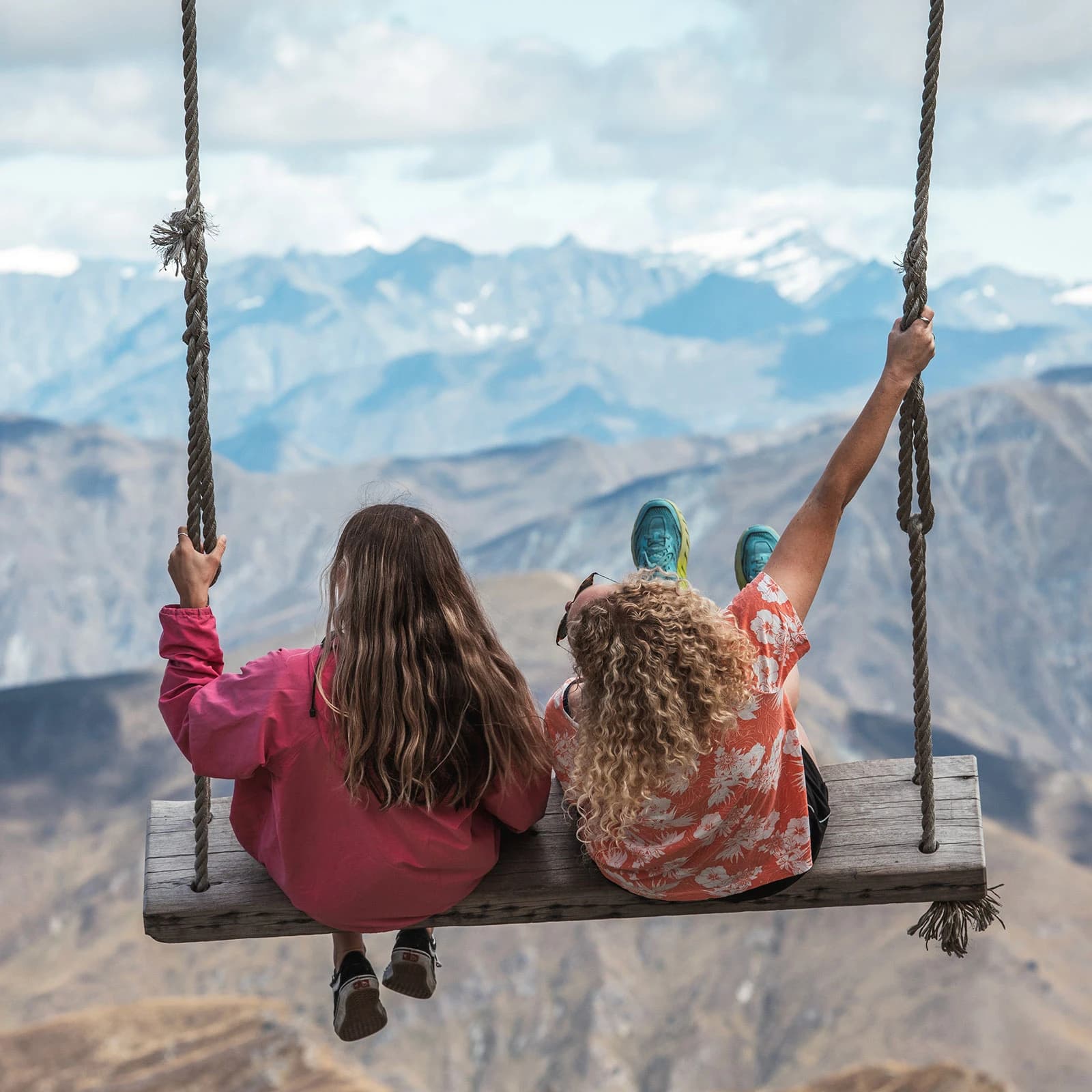 Two women sit on a large wooden rope swing facing away from the camera and swinging out over a background view of softly lit mountain ranges.