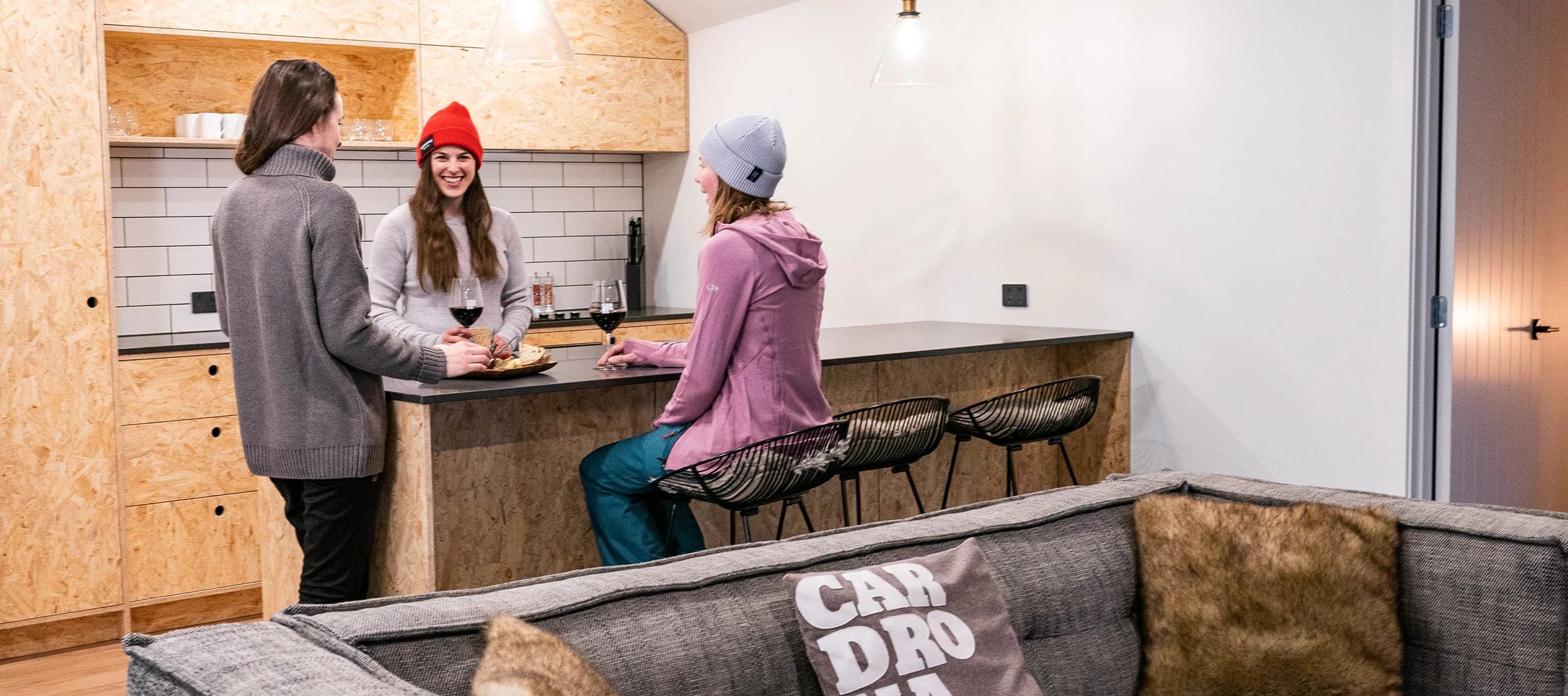 Three people gather around a modern kitchen counter in a cosy room and enjoy a glass of wine.