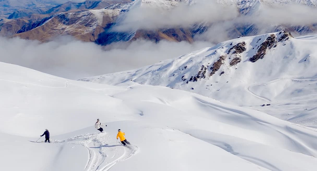 Three skiers gliding down a pristine, snow-covered mountain slope. The skiers are dressed in colourful gear—one in yellow, one in white, and one in black.