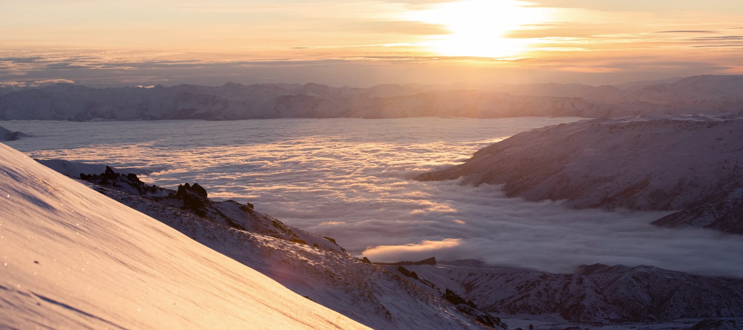 A breathtaking sunset over snow-covered mountains, with a sea of clouds filling the valleys below. The golden light of the sun casts a warm glow across the landscape, highlighting the ridges and creating a serene and majestic atmosphere.