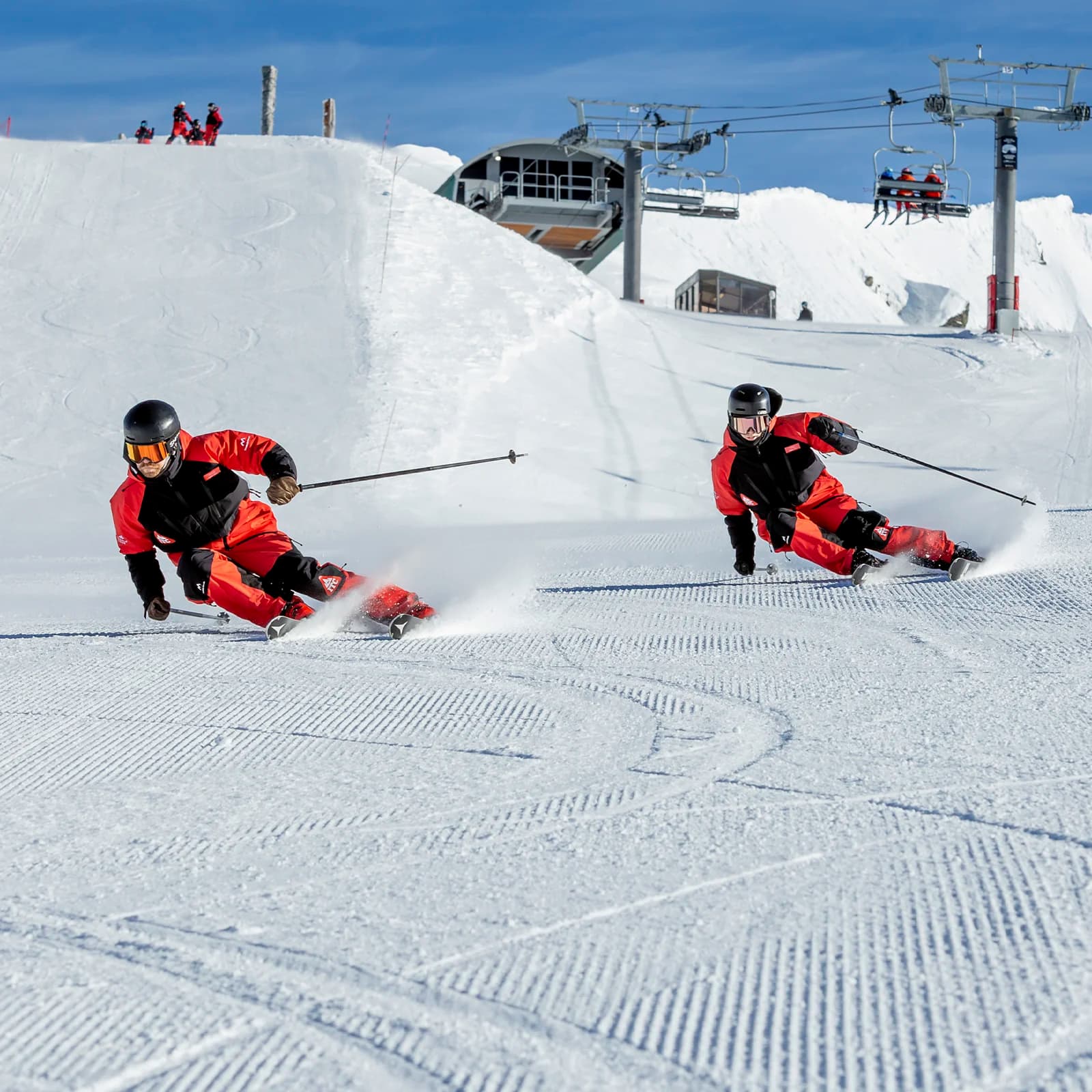 Two skiers in red and black outfits race down a freshly groomed slope, leaning into their turns and kicking up snow as they carve, with a ski lift and snowy peak in the background.