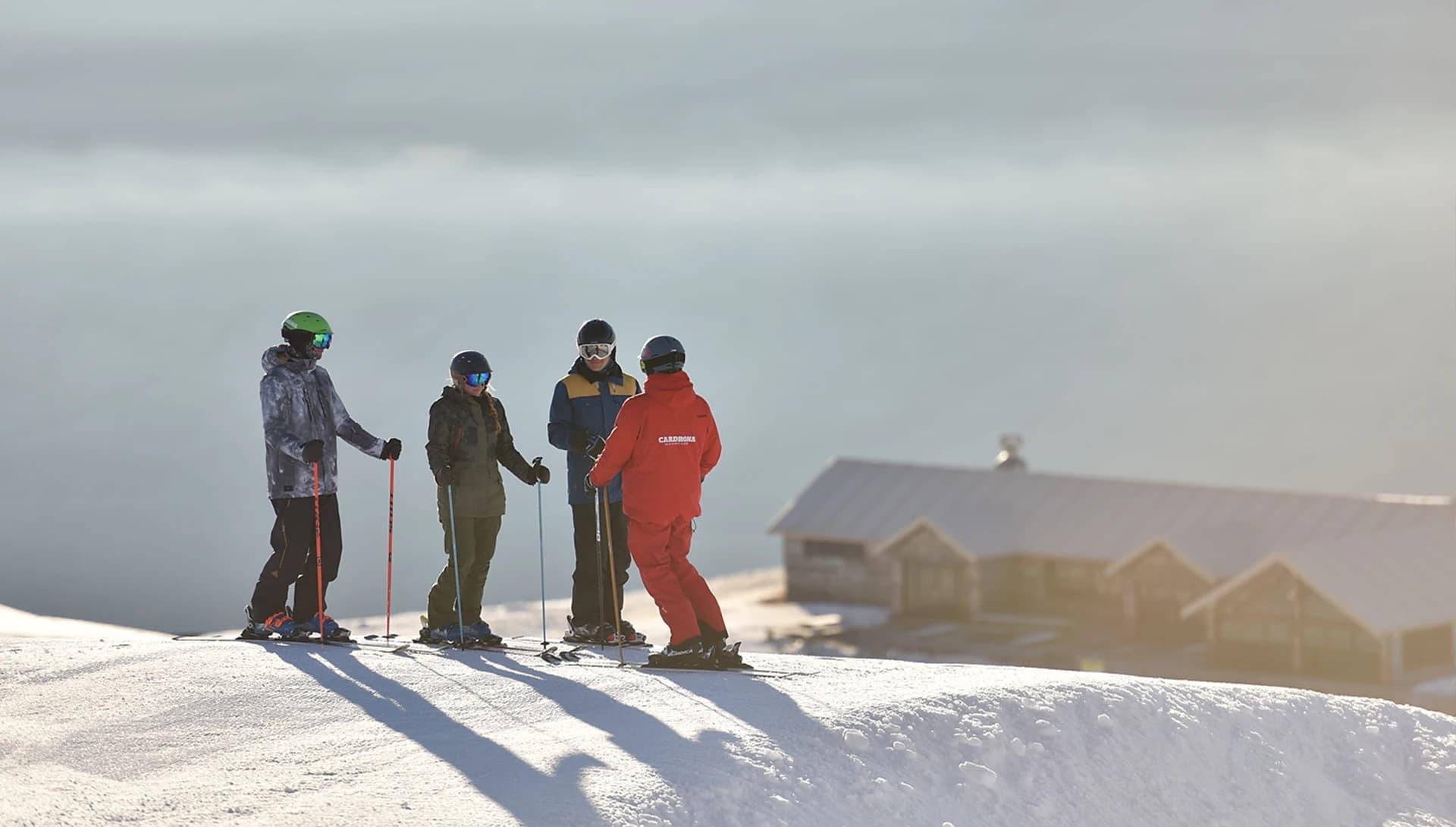 A ski instructor in a red jacket talks with three skiers on a snowy slope. The morning light creates long shadows, with a mountain lodge visible in the background under a serene sky.