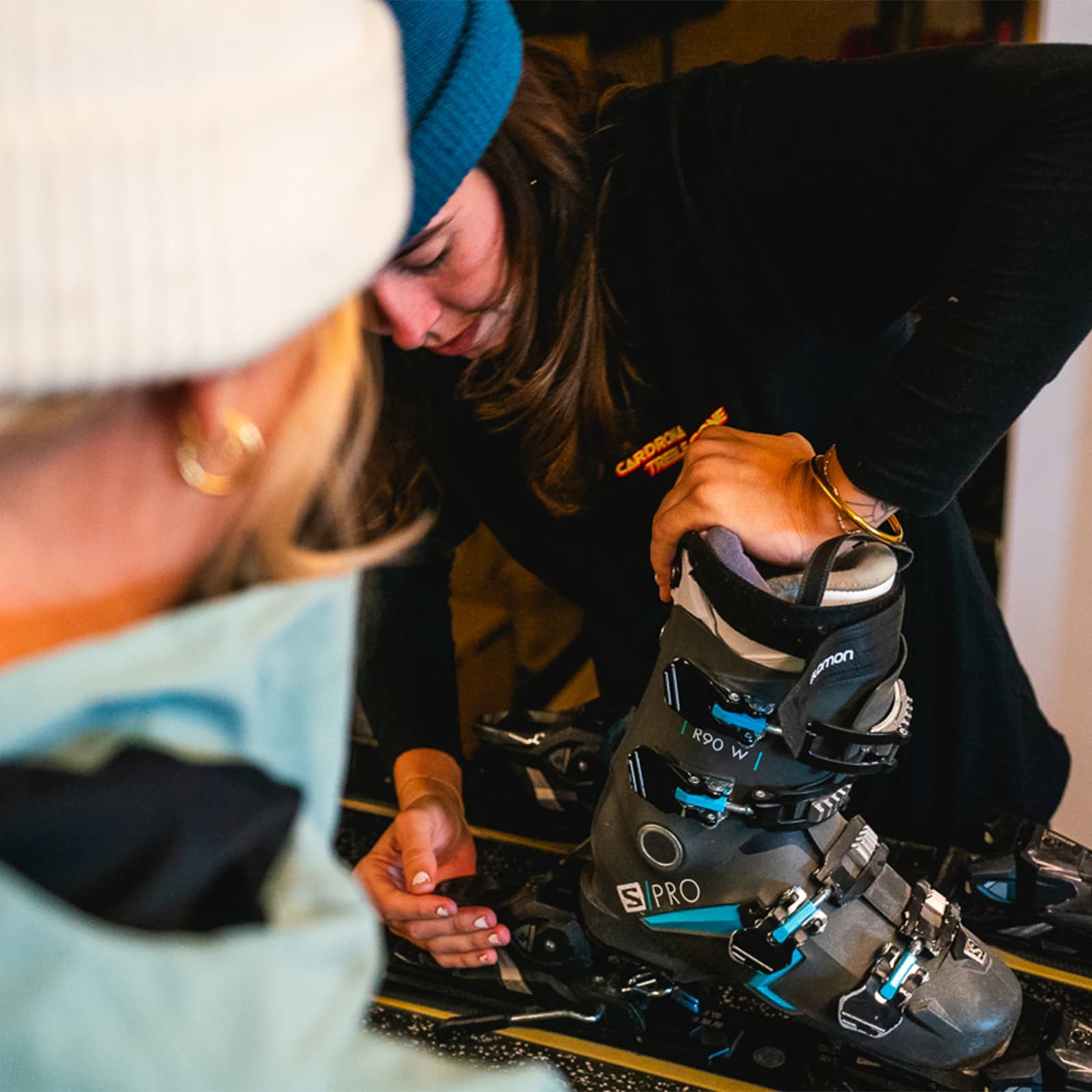 A ski rental technician adjusts a ski boot on a set of skis while a customer watches closely, both wearing winter hats in a cozy indoor setting.