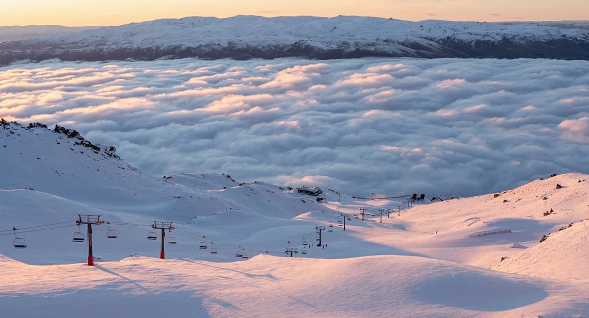 A view of Captains Basin at Cardrona Alpine Resort during sunrise, with soft pink light illuminating the snow-covered slopes and a sea of clouds below. Chairlifts are visible along with distant mountains in the background.