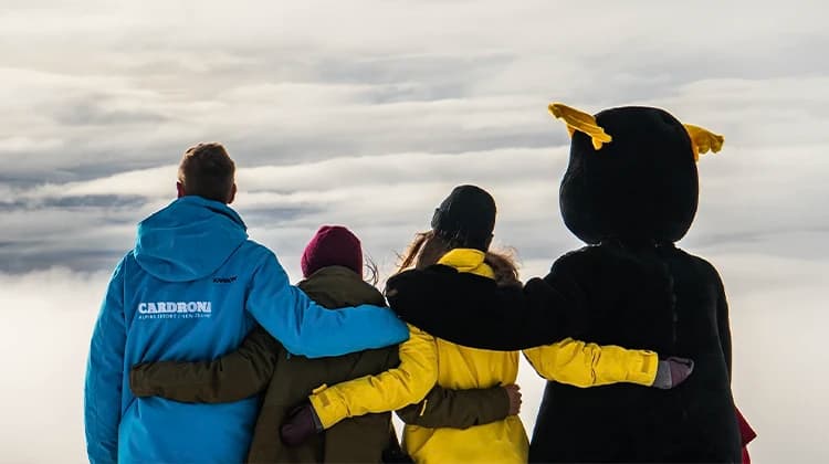 Three staff members and a mascot hold each other and look out at a stunning mountainous view through the clouds.