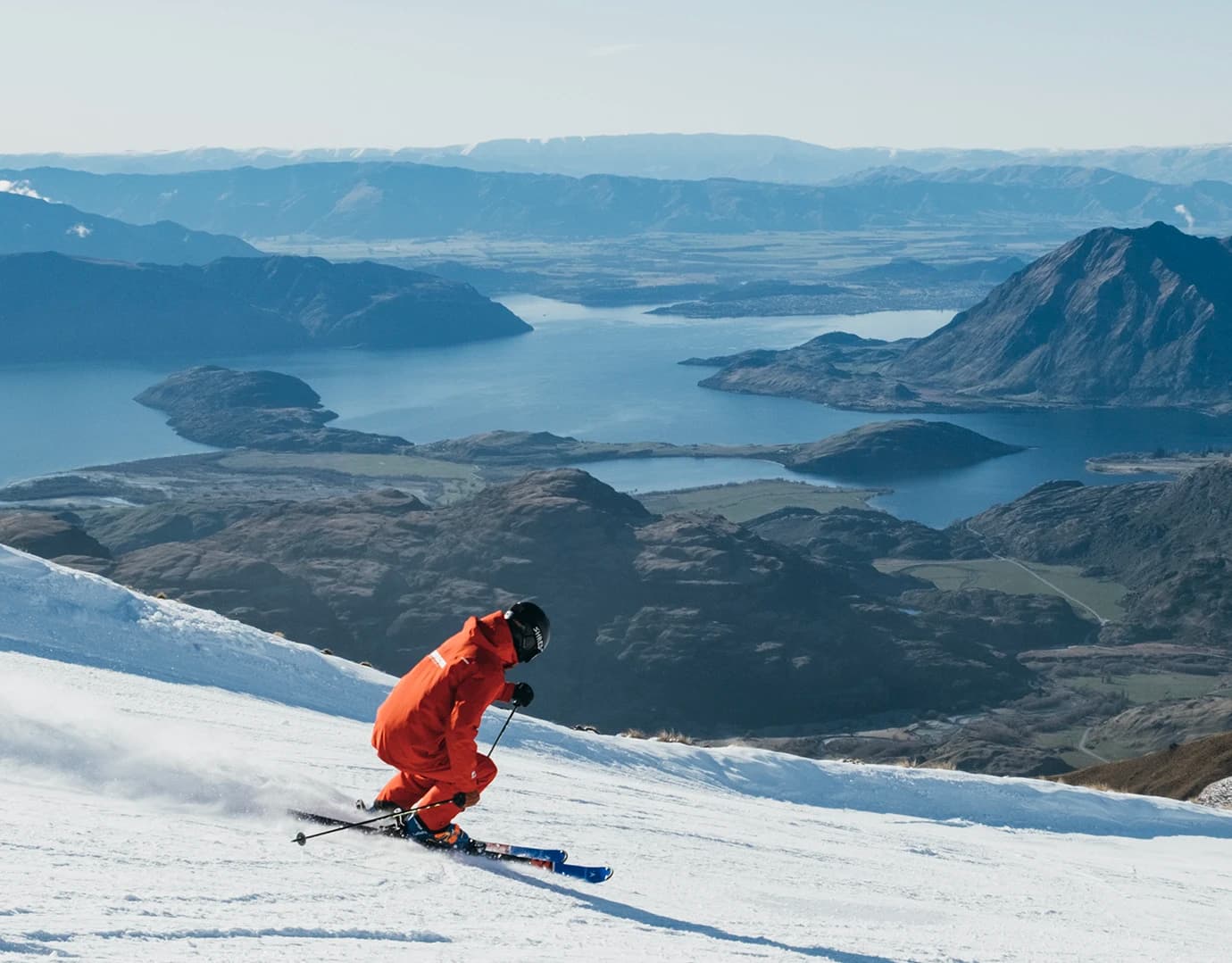 A skier in red glides down a snowy slope, leaving a trail of snow. In the background, a vast lake and mountains stretch under clear skies.