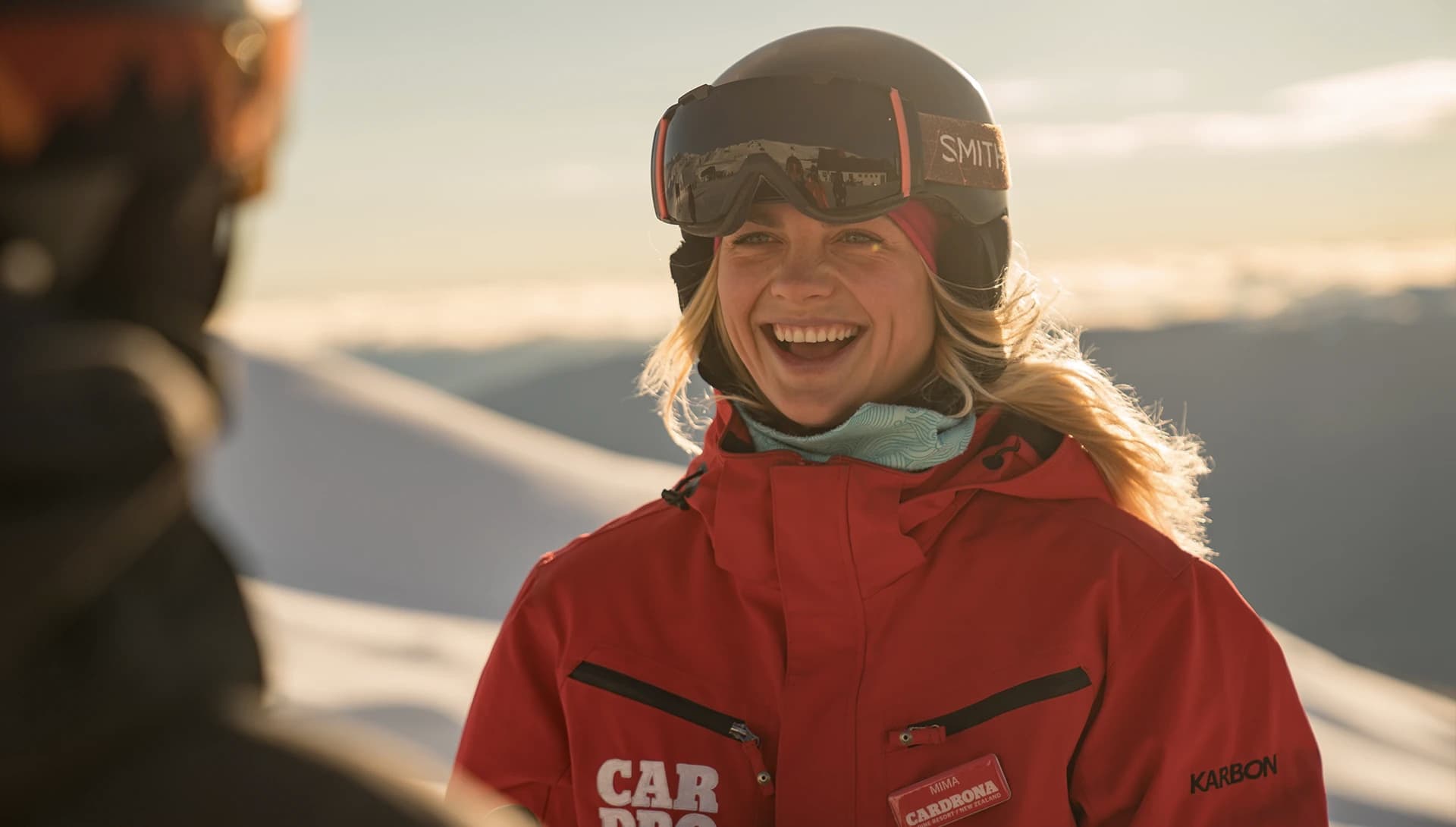 Close up shot of a woman smiling while standing outside with snowy mountains in the background. She is wearing a red ski jacket branded 'Cardrona' and a a black ski helmet with goggles branded 'Smith' on the helmet. 