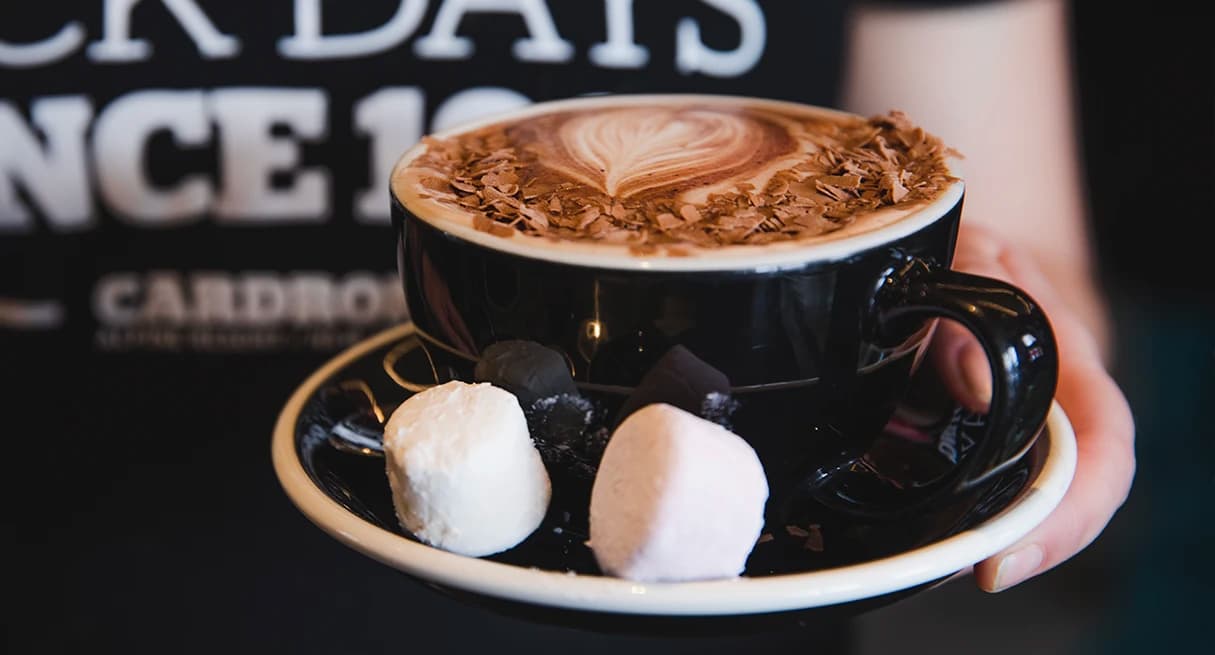 A person holds a hot chocolate with a pink and white marshmallow on the side.