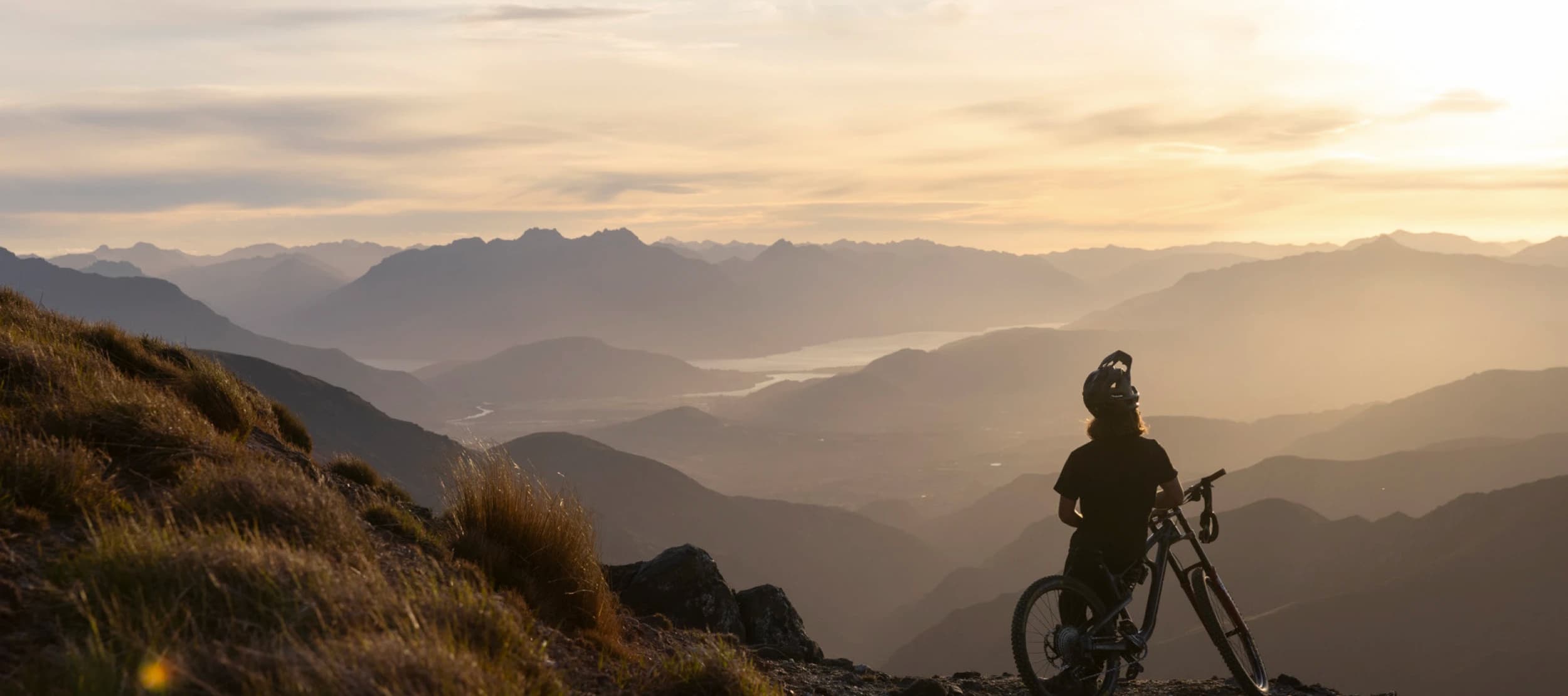 A mountain biker stands with their bike at sunset, overlooking the expansive Cardrona mountain range and valleys below.