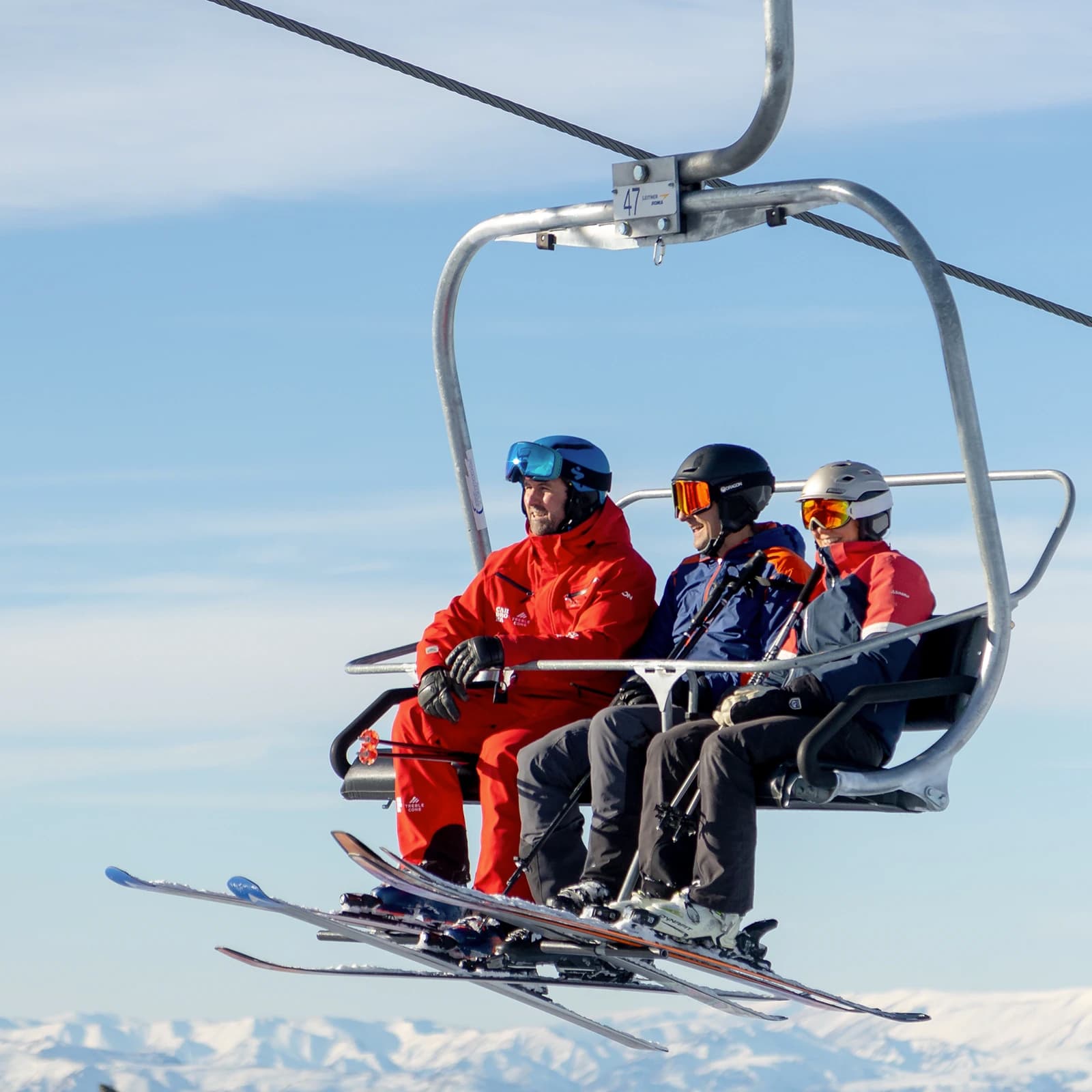 Three skiers sit smiling on a chairlift on a blue sunny day. One of the skiers, dressed in a bright red ski suit branded with 'Cardrona' is leading the other two in a ski lesson at Cardrona Alpine Resort.